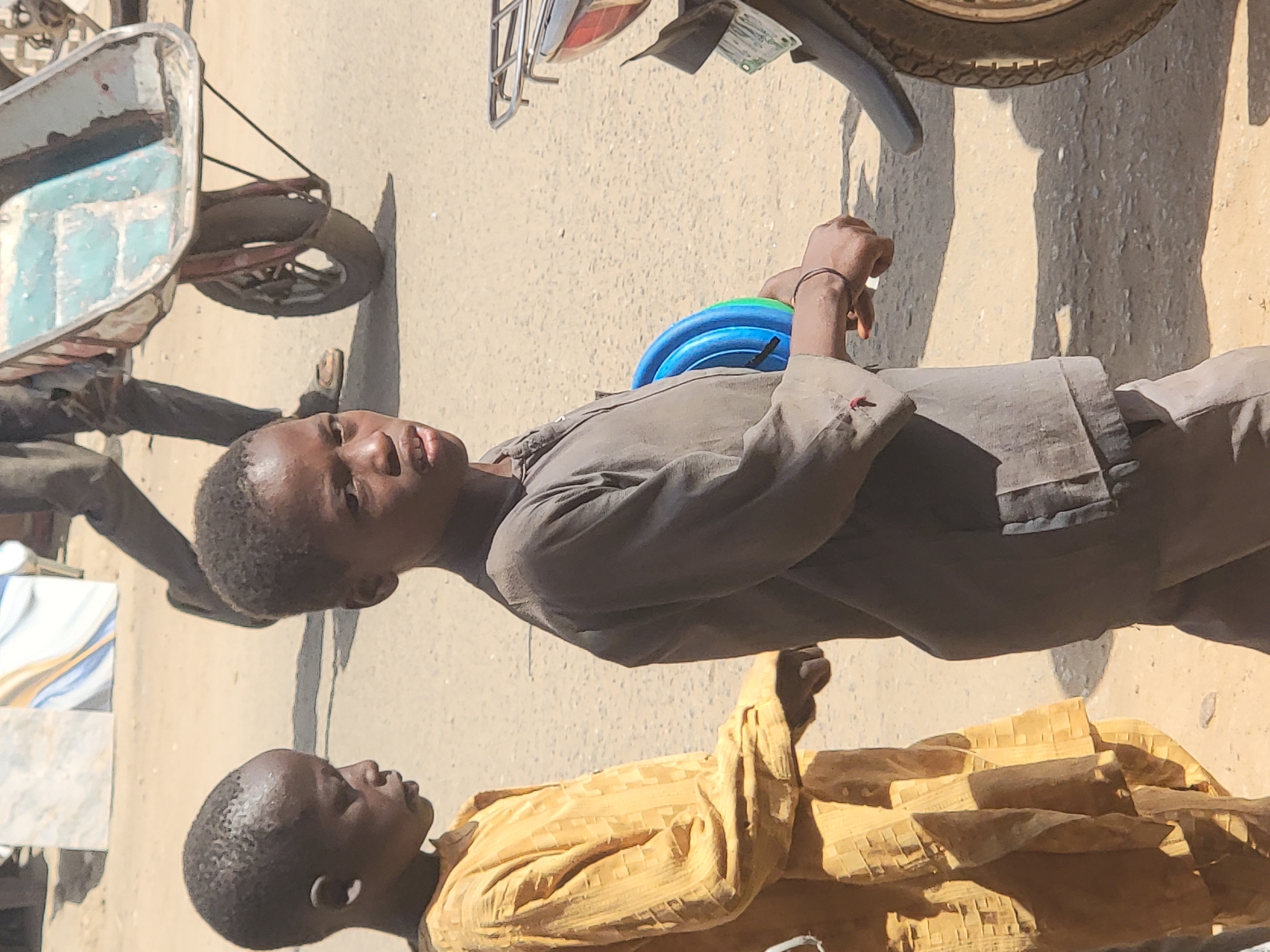 Two African children are in an outdoor setting. One child is wearing a yellow, patterned shirt and the other is wearing a grey button-down shirt. The child in the grey shirt is carrying a set of blue and green plastic rings. A wheelbarrow and the wheel of a motorcycle are also visible in the frame. The setting appears to be a street in Maiburiji, Nigeria.