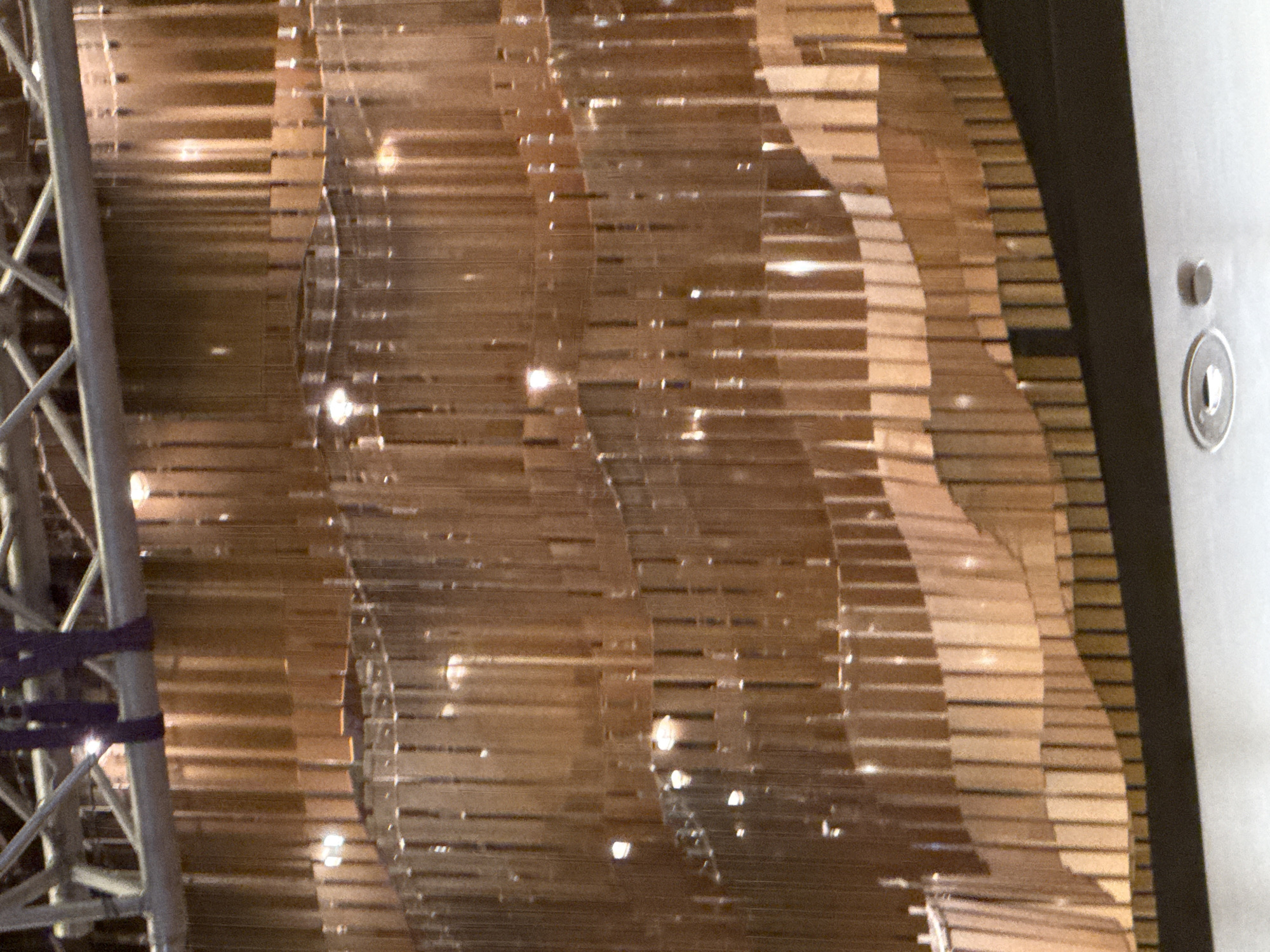 The image shows a close-up view of a decorative wall or partition made of many vertical, thin, wooden or metallic elements. These elements appear to be suspended and are arranged in a staggered, wave-like pattern, creating a sense of depth and texture. Lights are visible reflecting off the surfaces, suggesting an indoor setting. In the upper portion of the image, structural elements, possibly part of a ceiling or rigging system, are visible, along with some dark purple straps. The bottom of the image shows a dark, flat surface, possibly a stage or floor, and two circular, recessed fixtures. The overall impression is of a modern, artistic interior design feature. This image is from the Heydar Aliyev Center in Sabail Raion, Azerbaijan.
