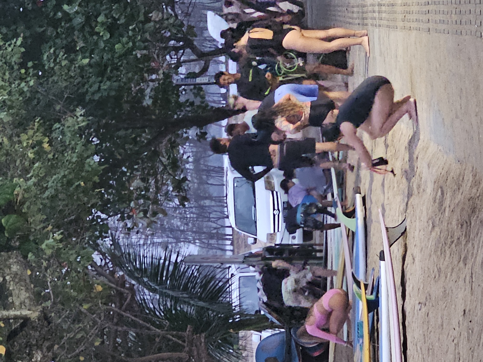 A group of people are gathered on a sandy area with surfboards scattered on the ground. Several white vans are parked in the background, and trees with dense foliage line the area. One person in the foreground is wearing a pink bikini and crouching near the surfboards. Another person in a black swimsuit is squatting on a paved surface, holding a leash in their hands. The scene appears to be a beachside location in Denpasar, Indonesia, possibly during a surfing event or lesson.