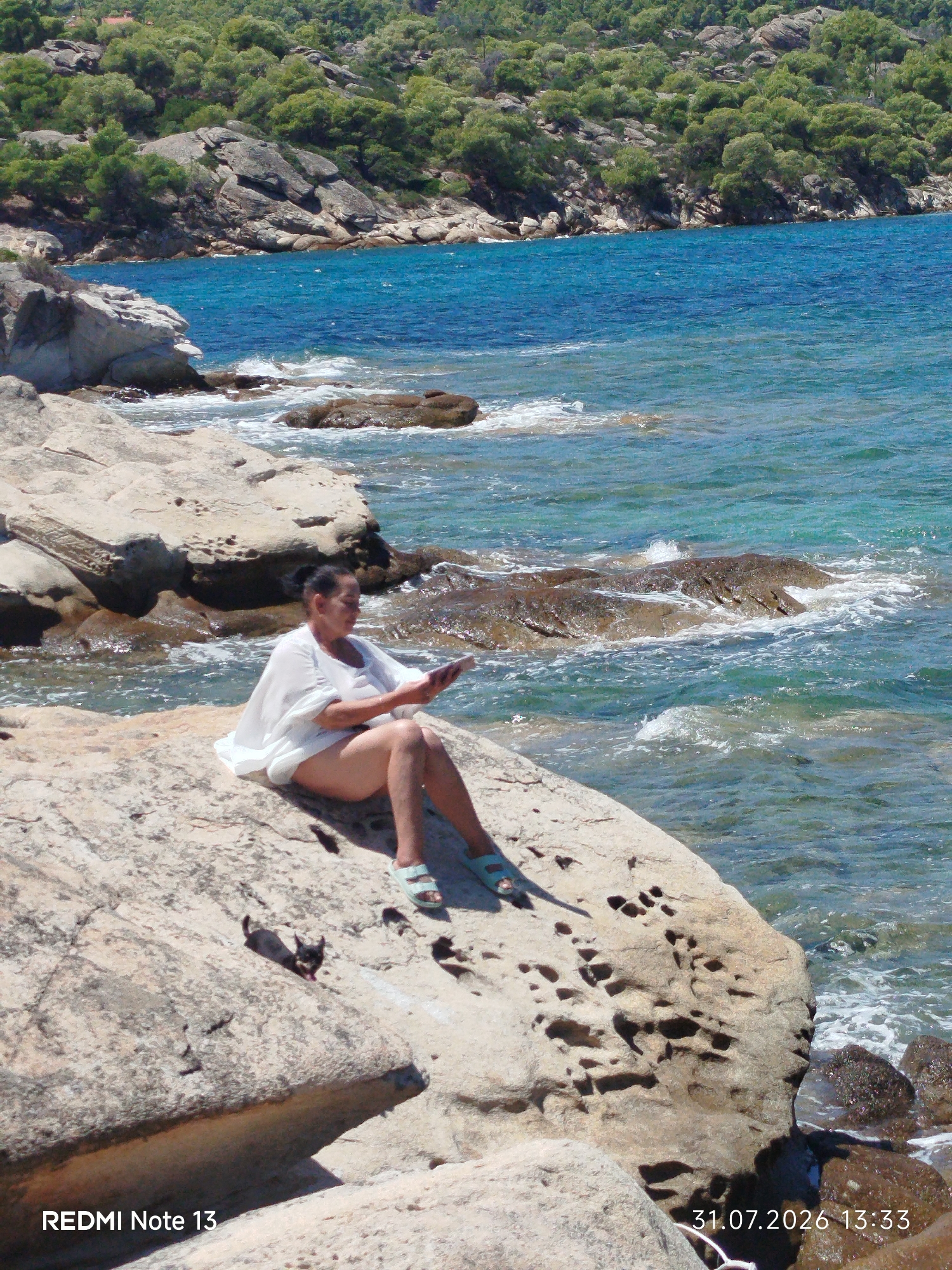 A woman is sitting on a large rock near the sea, holding a mobile phone. The water is blue and clear, with waves crashing on the rocks. The background shows a green, forested hillside. The image appears to have been taken in Sithonia Municipal Unit, Greece, as indicated by the timestamp and device information.
