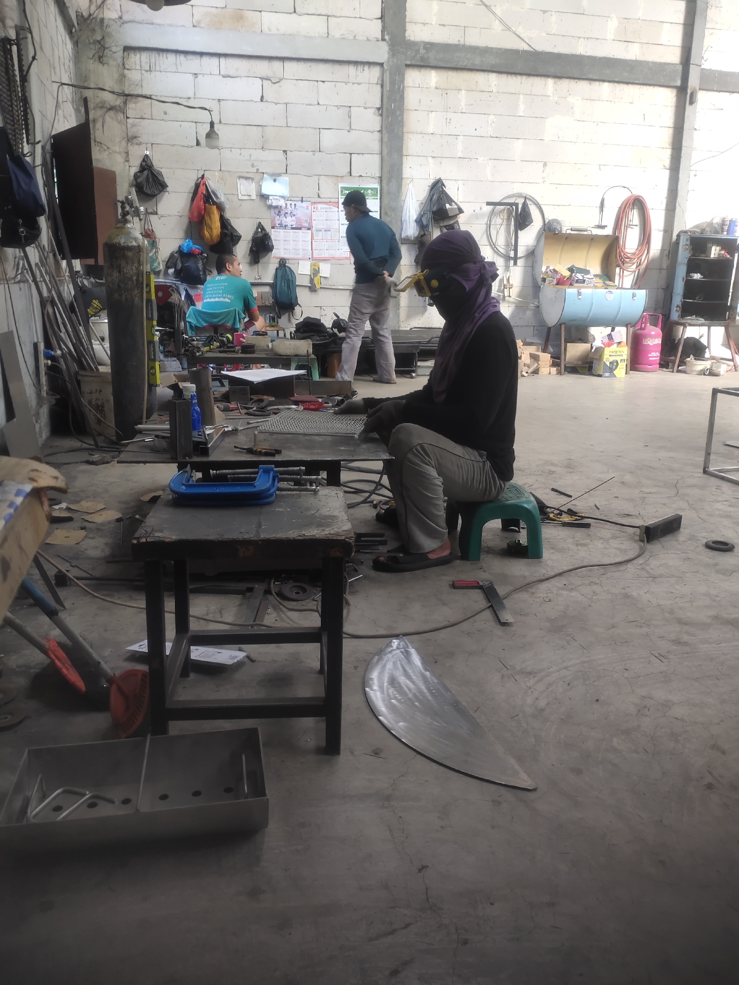 The image depicts an interior scene of a workshop, likely in Pataruman, Indonesia. The workshop appears to be a metal fabrication or welding facility, given the presence of welding equipment, metal stock, and unfinished metal projects.
In the foreground, a person wearing a dark long-sleeved shirt, grey pants, and a purple head covering with goggles is seated on a green plastic stool. This individual appears to be engaged in a task related to metalwork, possibly welding or grinding, as indicated by the protective gear. Welding equipment, including cables and a torch, are visible on the floor around them.
Several worktables are present in the scene. One table in the foreground holds a large blue C-clamp. Other tables are cluttered with tools, metal pieces, and what appears to be a roll of wire mesh. In the background, two other individuals are visible. One, wearing a teal shirt, is seated at a table. Another, in a blue shirt and grey pants, stands and appears to be observing or interacting with the person in the teal shirt.
The workshop environment is utilitarian, with cinder block walls and a concrete floor. Various tools and materials are stored on shelves and against the walls, including gas cylinders, hoses, bags, and metal rods. A large, curved piece of metal, possibly a component for a larger structure, lies on the floor in the mid-ground. The lighting in the workshop is somewhat dim, with light sources appearing to be overhead lamps and possibly natural light from unseen openings. The overall impression is of a functional workspace where metal fabrication or repair is actively taking place.