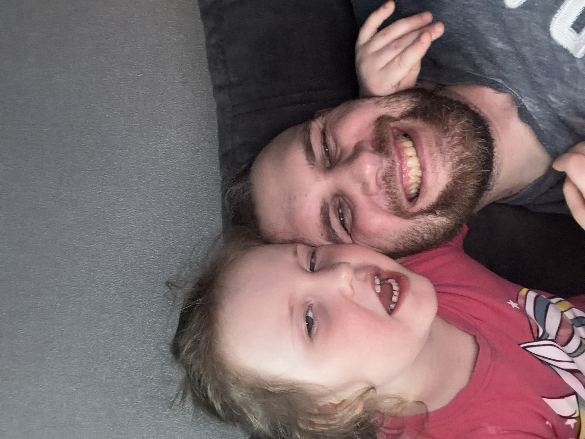 A man and a young child are lying on a gray surface. The man has a beard and is wearing a gray shirt. The child is wearing a pink shirt with a unicorn on it. Both are smiling and appear to be in a relaxed, playful mood. There is a dark gray pillow or cushion behind the man's head. The location is unknown. The image does not show any identifiable features that would indicate the location is Chaikovsky, Russia.