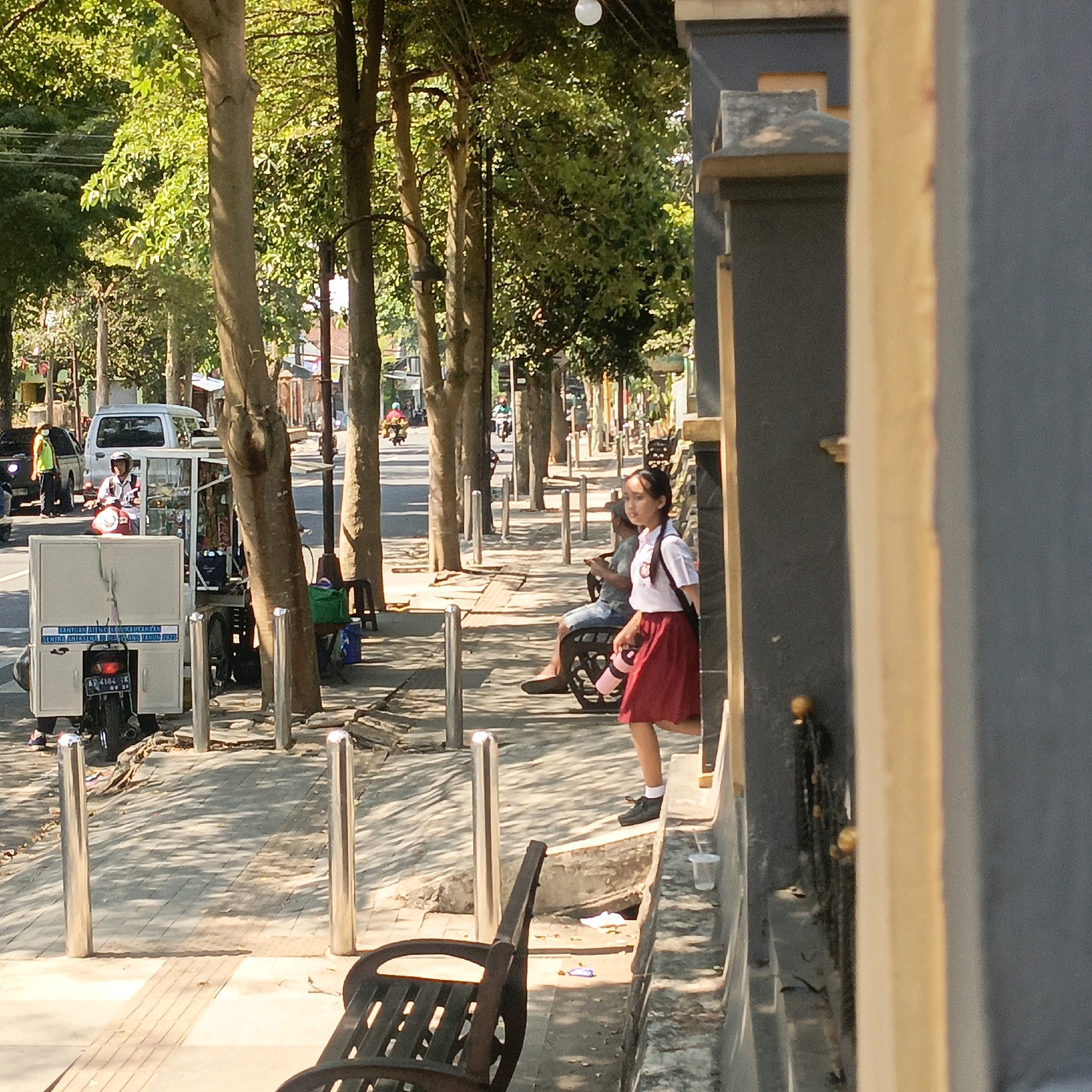 This image depicts a street scene in Salatiga, Indonesia, under bright daylight. In the foreground, a young girl wearing a school uniform, consisting of a white shirt and a red skirt, walks along a paved sidewalk. She is carrying a pink water bottle and a black bag. To her left, a park bench is visible, and further down the sidewalk, another person is seated on a bench, holding a mobile phone. The sidewalk is lined with bollards and trees casting dappled shadows. Beyond the sidewalk, a street is visible with traffic, including a silver car and several motorcycles. A food cart, equipped with a refrigerator unit and a motorcycle for mobility, is parked on the side of the road. The background shows more trees and buildings. The overall atmosphere is that of a typical urban environment in Indonesia, with signs of daily life and commerce.