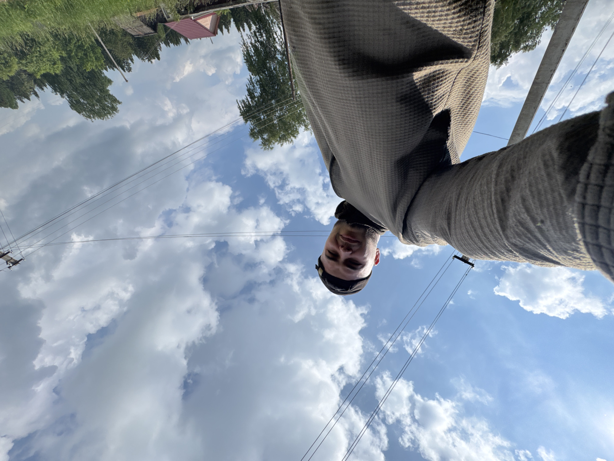 A low-angle, wide-shot selfie is taken from the perspective of a person holding a camera with their left arm extended. The subject, a young man wearing a dark baseball cap and a textured, dark gray long-sleeved shirt, is positioned in the lower left quadrant of the frame, looking towards the camera. His face is partially obscured by the angle of the shot.
The background consists of a bright blue sky with scattered, fluffy white clouds. Overhead power lines crisscross the upper portion of the image. Below the sky, a rural landscape is visible, characterized by a concrete utility pole on the left, dense green trees, and a glimpse of rooftops from houses in the distance. The ground is covered in tall green grass. The scene suggests an outdoor setting in a rural or semi-rural area. The location is identified as Borodulino, Russia.