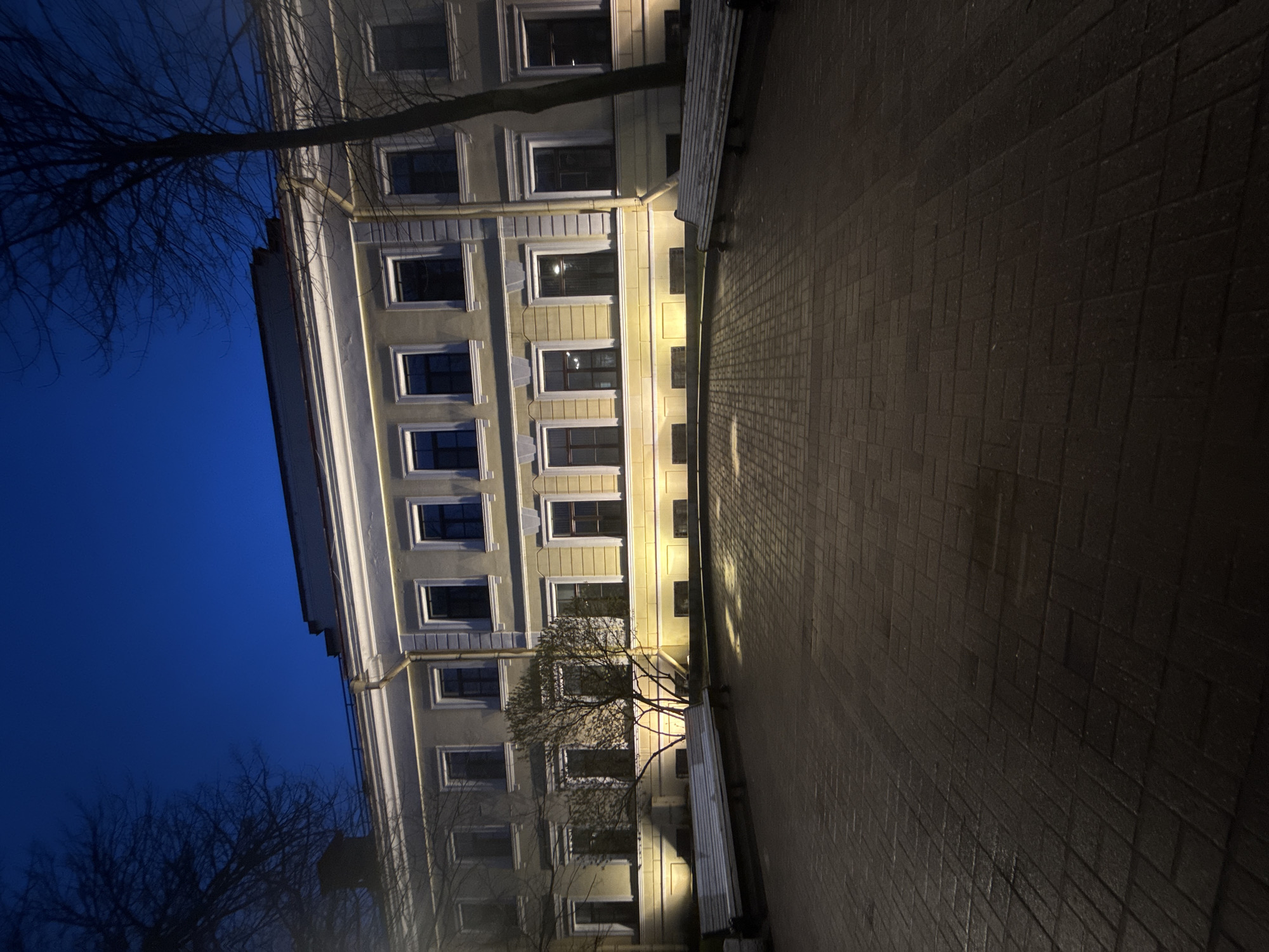 The image depicts a building in Saint Petersburg, Russia, at night. The building has multiple stories, with rows of windows, and is illuminated by artificial lighting. The building’s facade is visible, along with the adjacent brick-paved sidewalk. A tree is partially visible in the foreground, adding to the urban landscape. The sky is dark blue.