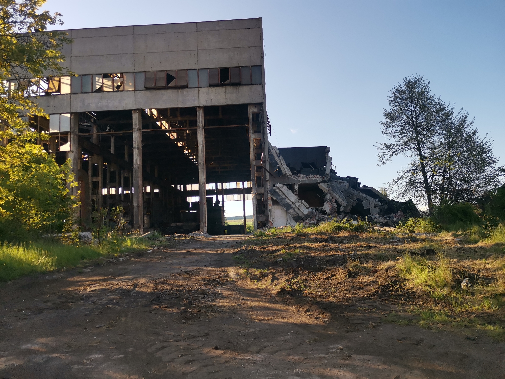 The image shows an abandoned and partially destroyed industrial building located in what appears to be Zhivkovo, Bulgaria. The building is a large, concrete structure with a significant portion of its roof and walls collapsed into a pile of rubble next to it. The remaining portion of the building features a framework of concrete columns and beams, with missing windows and visible interior elements. A dirt road leads up to the building, with patches of grass and vegetation growing on either side. In the background, a tree stands near the wreckage. The sky is clear and blue.