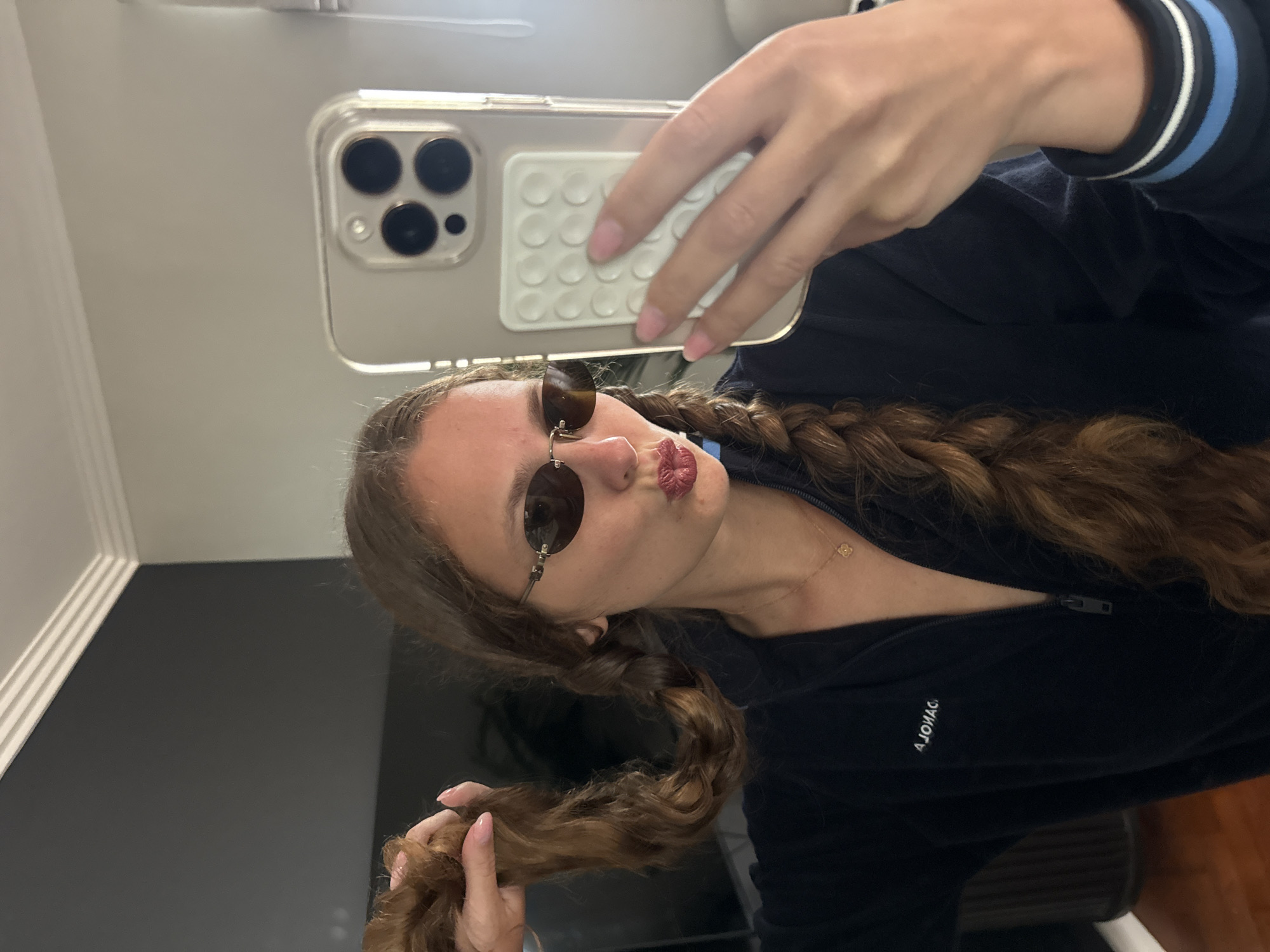 A young woman is taking a selfie indoors, holding an iPhone in her right hand. She has long, brown hair styled in braids and is wearing round sunglasses. She is pouting her lips towards the camera. The woman is wearing a dark blue jacket, and a necklace is visible around her neck. The background appears to be a room with a gray wall and white molding. There is no indication of the city of Cape Town in this image.
