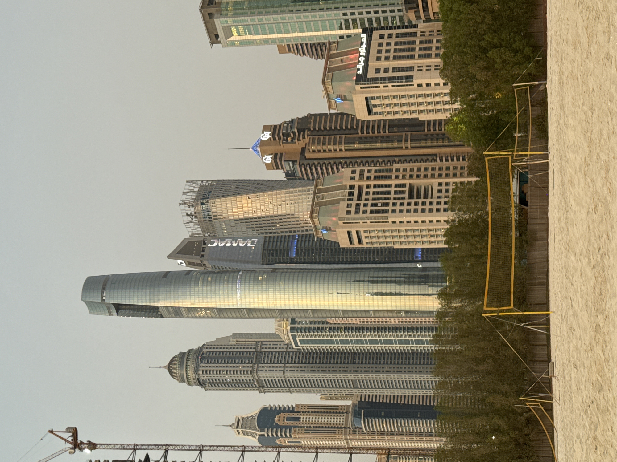 The image shows a beach with a volleyball net in the foreground, set against a backdrop of skyscrapers in Dubai, United Arab Emirates. The sand is light brown, and the volleyball net is yellow with support poles. Behind the net, a line of dense green trees separates the beach from the towering buildings. The skyscrapers are modern and vary in design and height, reflecting the city's architectural style. Some buildings have distinct lighting features, and a construction crane is visible on the far left of the frame. The sky is a hazy, light gray, suggesting it might be dawn, dusk, or an overcast day. The scene captures a blend of recreational space and the dense urban landscape characteristic of Dubai Marina. There are no people or animals visible in the image.