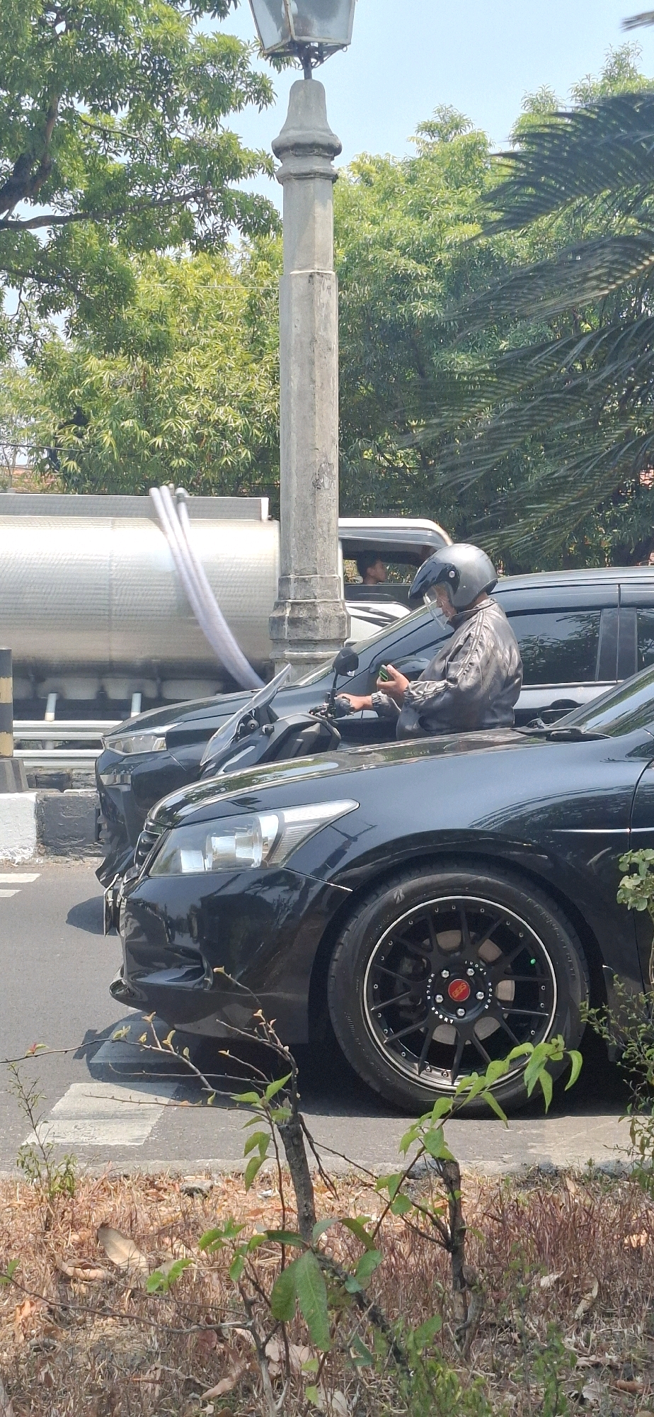 A motorcycle rider wearing a black helmet and a black jacket is looking at a smartphone while stopped in traffic in the city of Surakarta, Indonesia. The rider is seated on a black motorcycle, which is positioned in front of a black car. Behind the motorcycle and car, a large silver tanker truck is visible, with steam or exhaust emitting from pipes on its side. A street lamp post stands to the right of the tanker truck. The scene appears to be a roadside during daytime, with trees and vegetation visible in the background and foreground. The black car has a custom wheel with a red and black center cap. The ground in the foreground is covered with dry grass and small branches.