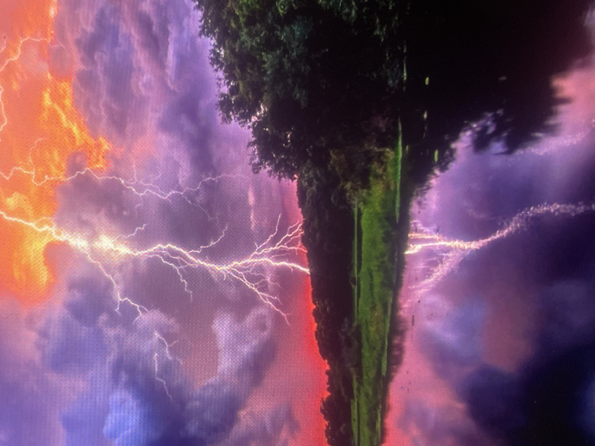 The image depicts a dramatic stormy landscape located in the city of Zelenyi Hai, Ukraine. A prominent, intensely bright white-yellow lightning bolt descends from the upper right quadrant of the sky, striking towards the horizon on the left. The sky exhibits a vivid and saturated color palette: fiery orange and red hues are concentrated towards the upper right and the distant horizon, gradually transitioning into deep purples, blues, and grays within dense cloud formations across the central and left portions. A dark, dense tree line forms the horizon, silhouetted against the bright red sky. Below the tree line, a strip of vibrant green land is visible. The foreground is dominated by a body of water, likely a river or lake, which mirrors the dramatic sky in shades of purple and pink, clearly reflecting the shimmering lightning bolt. No human subjects, animals, or man-made structures are overtly visible within the frame.