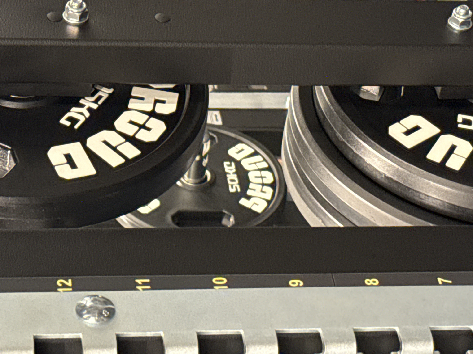 The image presents a close-up view of a weight rack, likely within a fitness facility. The rack is constructed from dark-colored metal, with visible numbered slots ranging from 7 to 12. A shiny metal fixture is affixed near the number 12.
Several weight plates are positioned on the rack. These plates are black with white lettering. The plates are identified as 15KG and 5OKG. The "SOHC" brand is also printed on the plates.
The location of this image in Plovdiv, Bulgaria, cannot be definitively determined from the provided visual information. Without additional context or identifying elements within the scene, specifying its exact location in the city is not possible.