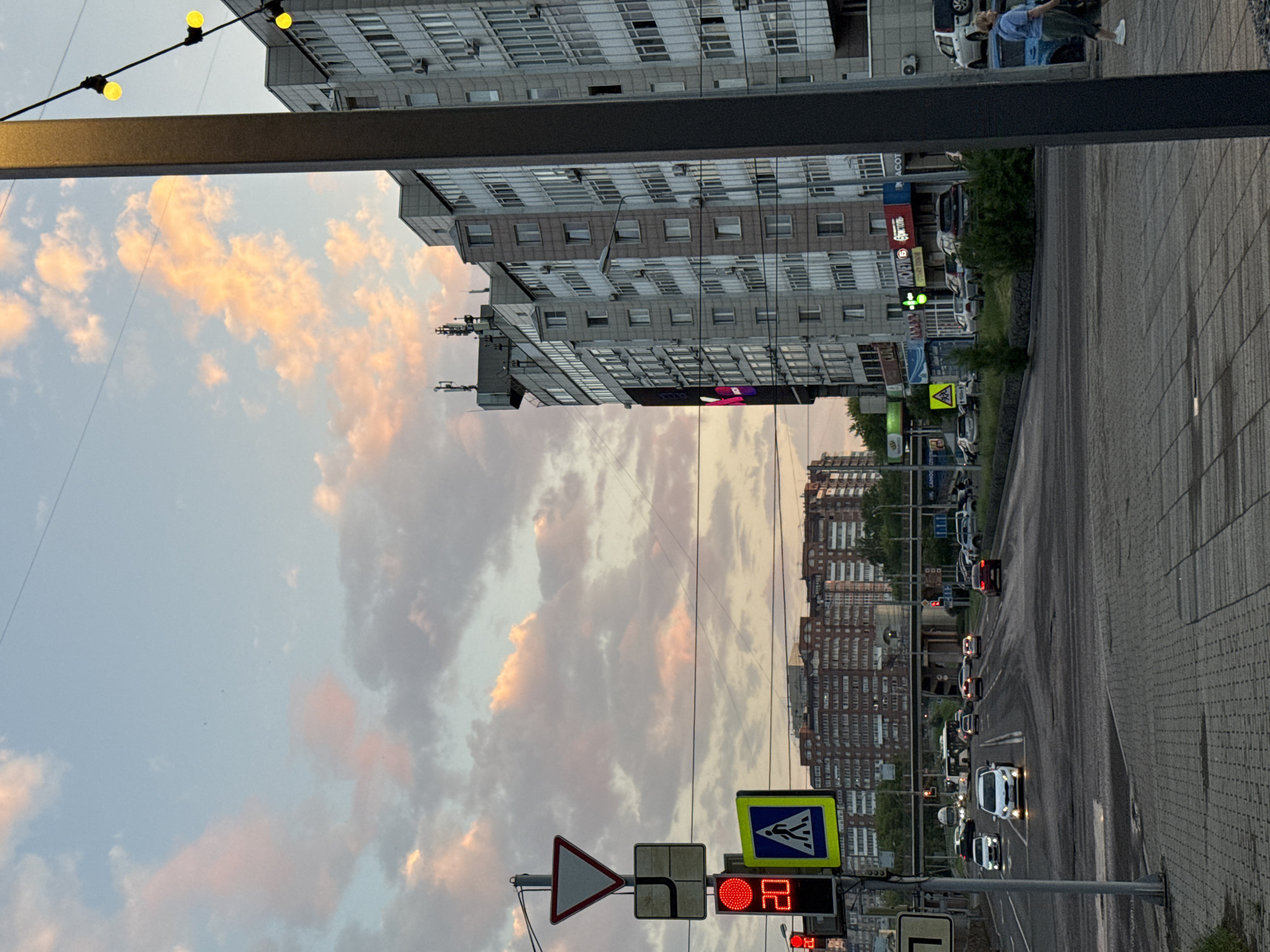 The image depicts an urban street scene at dusk, with a pink and orange-hued sky filled with clouds. In the foreground, a pedestrian crossing is visible, marked by a yellow and blue sign and a traffic light showing a red signal with the number "20" displayed below it, likely indicating a countdown. A large grey pole stands to the right, adorned with decorative yellow string lights.
Further down the street, several multi-story apartment buildings line the right side of the road, featuring numerous windows and air conditioning units. A large digital billboard is affixed to one of these buildings. Cars are present on the road, some with headlights on, suggesting ongoing traffic. A white bus and other vehicles are visible in the distance, as is an overpass. On the left side of the image, more apartment buildings, some with balconies, are seen. The road surface appears wet in places, and the sidewalks are paved with grey tiles. A woman in a blue shirt and dark pants walks along the sidewalk on the right.
The scene is consistent with an intersection in the city of Krasnoyarsk, Russia, likely near residential areas and commercial establishments. The presence of Cyrillic signage on the buildings, such as "ВИОЛЕТА" and "МАГНИТ", further supports this location. The overall atmosphere is that of a typical city evening commute or activity.