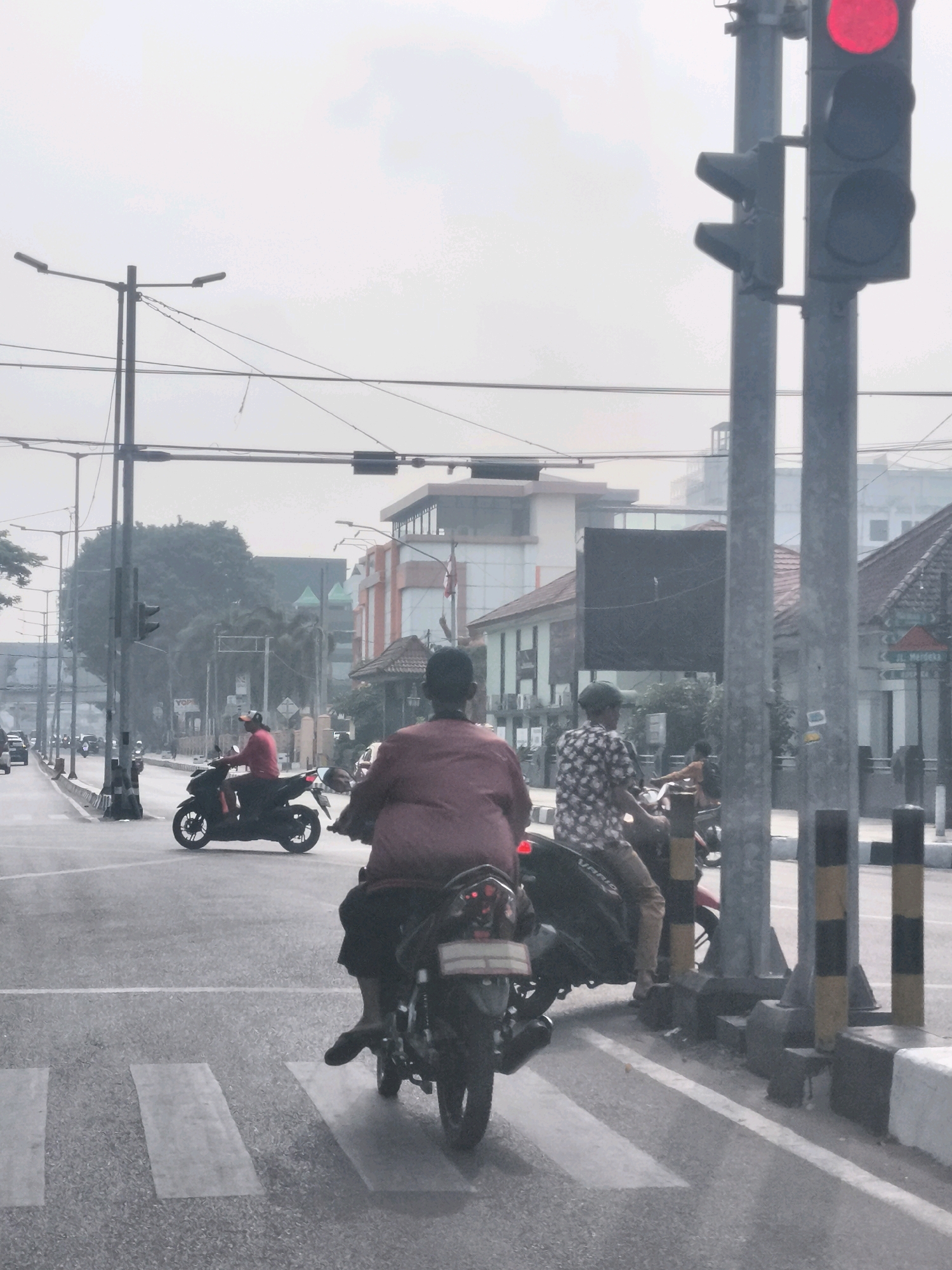The image depicts a traffic intersection in Palembang, Indonesia, on a hazy day. The foreground features a pedestrian crossing with two motorcycles stopped at the red light. On the left, a motorcycle with a rider in a maroon shirt and black pants is visible from the rear. On the right, another motorcycle with a rider in a patterned shirt and tan pants is also stopped. In the background, there are buildings with modern architecture, some appearing to be shops or offices. Traffic lights are visible, with the top light showing red. Lamp posts and overhead electrical wires are also present, contributing to the urban landscape. The overall atmosphere is that of a typical city street scene with ongoing traffic.