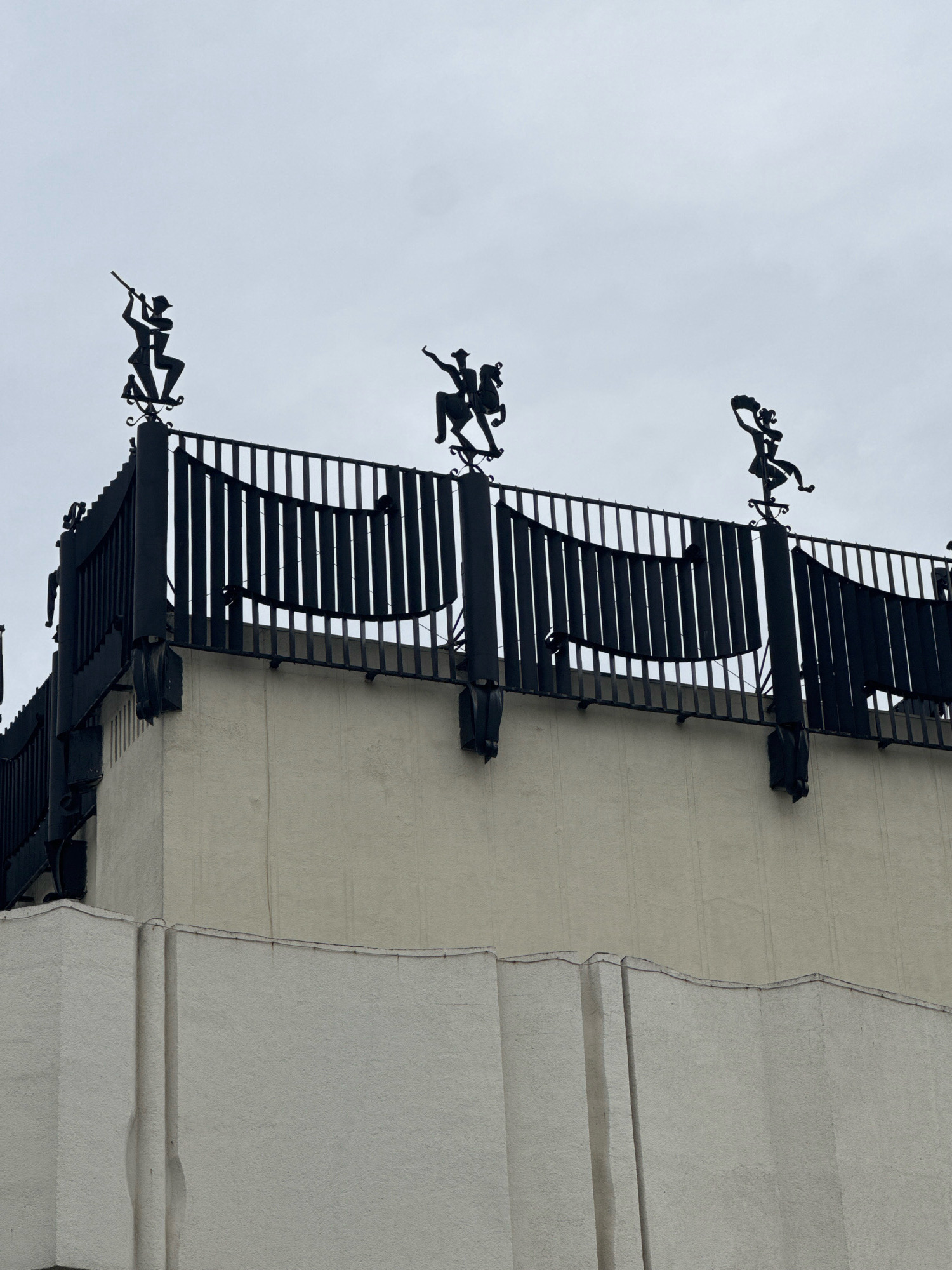 This is a low-angle exterior shot of the roofline of a building, likely in Ufa, Russia, under an overcast sky. The building's upper facade is a light cream color with a textured finish. A black metal railing runs along the edge of the roof. Attached to the railing are several decorative silhouetted figures, also in black metal.
From left to right, the figures depict:
1. A person in a dynamic pose, holding what appears to be a long staff or pole.
2. Two figures, one on horseback, with the rider's arms raised.
3. A lone figure in a dancing or celebratory pose, holding a piece of fabric or a flag above their head.
The railing itself has vertical bars with decorative scrolling elements between some of the figures. The building's lower facade, visible at the bottom of the image, is a brighter white with fluted vertical elements and a slightly wavy top edge. The overall impression is one of architectural ornamentation.