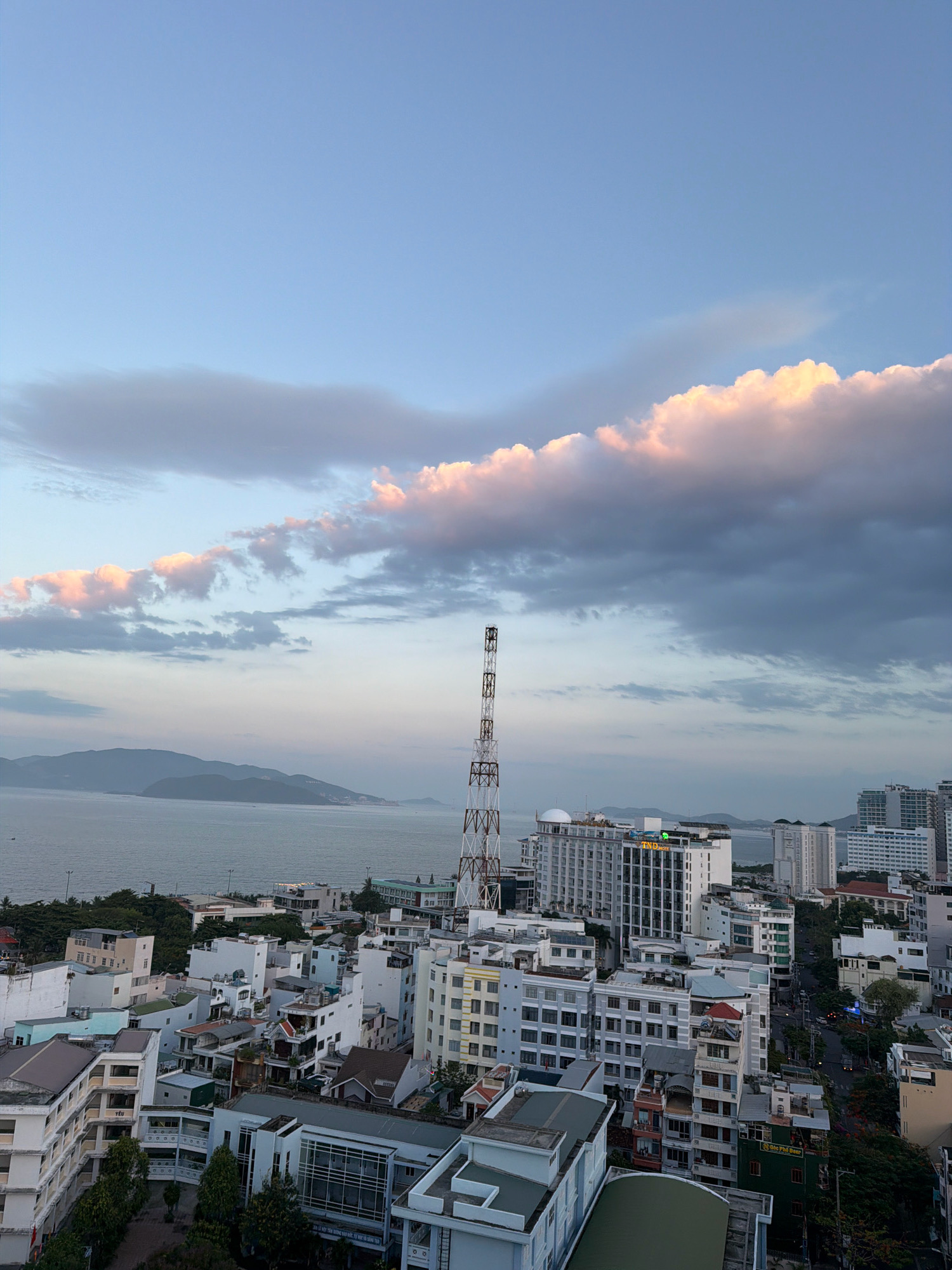 The image shows a cityscape of Nha Trang, Vietnam, with the ocean in the background. The sky is a pale blue with scattered clouds, some of which are tinged with pink from the setting sun. In the foreground, there is a dense collection of buildings of varying heights and architectural styles, indicative of an urban environment. A tall, lattice-like radio tower stands prominently in the mid-ground, near the coast. The ocean is calm, with distant islands or peninsulas visible on the horizon. The overall impression is a coastal city at dusk.