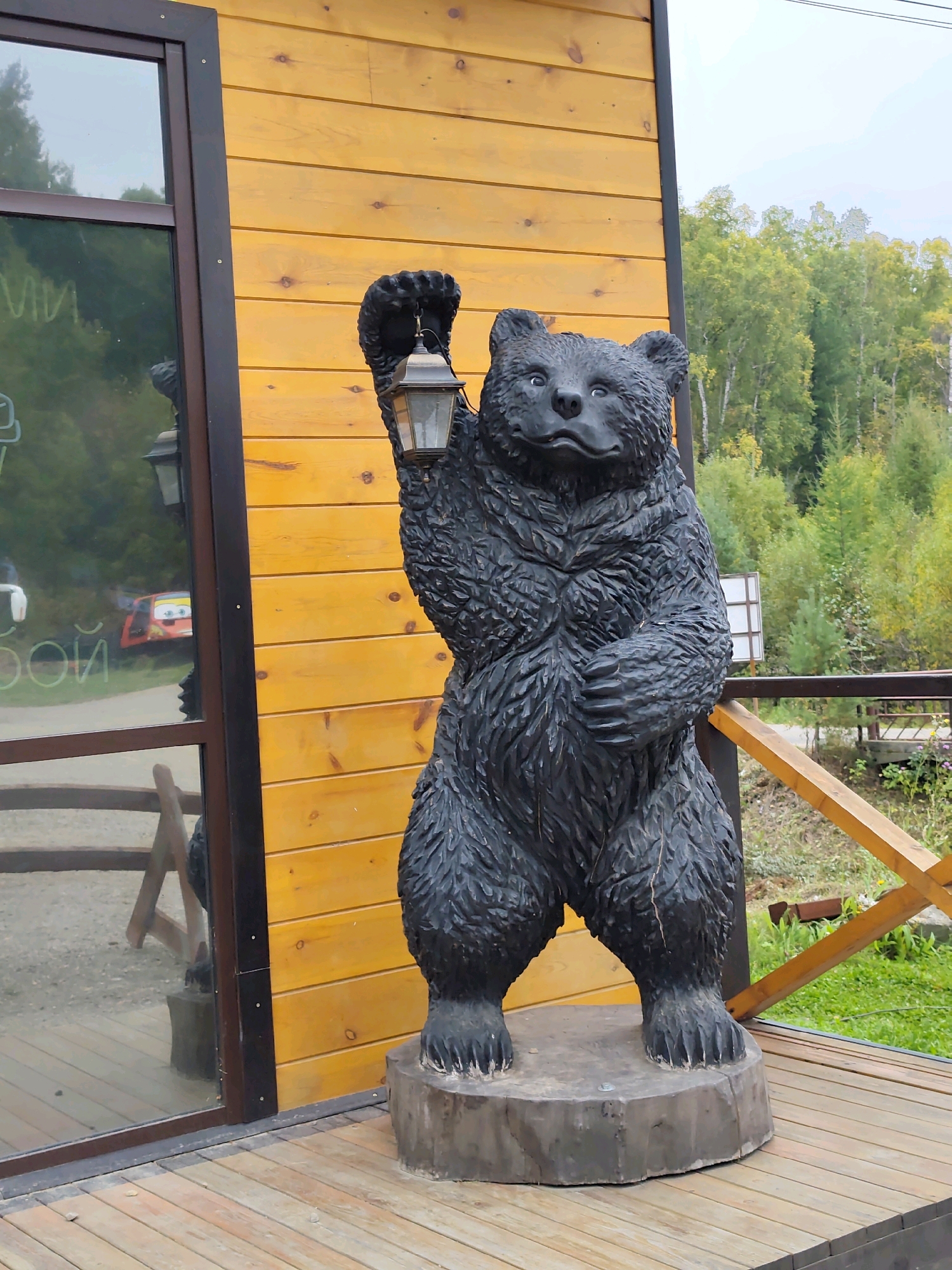 A life-sized, dark-colored, carved wooden statue of a bear stands on a wooden platform in front of a yellow wooden building. The bear is holding an old-fashioned lantern in its right paw, raised above its head. The statue is intricately carved to depict fur texture. The building has a large glass window on the left, reflecting trees and a red vehicle. A wooden railing with diagonal supports is visible behind the bear and to the right. The ground is covered in grass and a wooden walkway. The background shows a dense forest with green and yellow foliage, suggesting an outdoor location. The scene appears to be in the city of Listvyanka (Irkutsk Oblast), Russia.