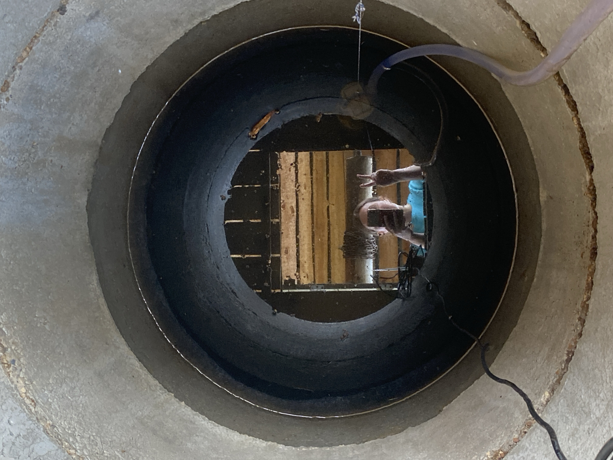 The image shows a view down a concrete well or large diameter pipe. At the bottom, wooden planks are visible, partially submerged in water. Hoses and electrical cords run down into the well. A human reflection is visible in the water, showing a person wearing blue clothing, possibly holding a phone or camera. The location is not discernible from this image alone.