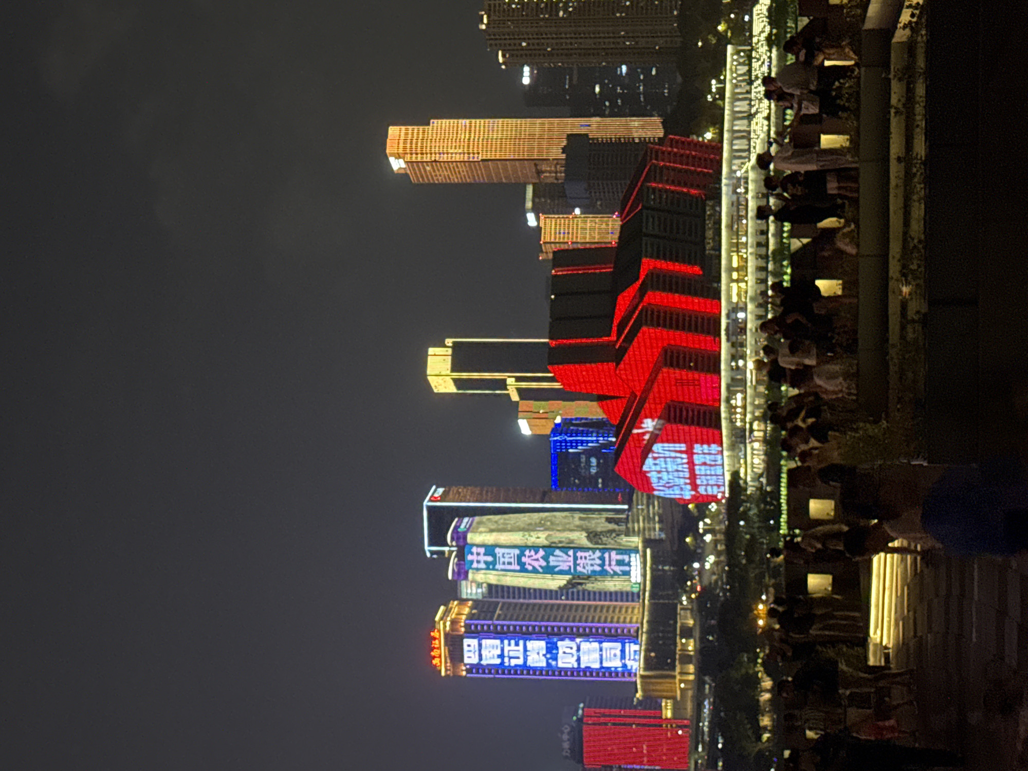 The image depicts a nighttime scene in Yuzhong District, Chongqing, China, showcasing a cityscape illuminated by vibrant lights. In the foreground, a group of people are gathered, some silhouetted against the bright lights of the buildings. They appear to be standing on an elevated walkway or viewing area, looking towards the cityscape. Behind them, several modern skyscrapers rise into the dark sky, adorned with colorful LED displays. One prominent building displays Chinese characters in bright blue, reading "中国农业银行" (Agricultural Bank of China), and another displays characters in white and purple, which are partially obscured. Further to the left, a red building is also illuminated with lights. The buildings suggest a bustling urban environment, likely a commercial or financial district, with a promenade or park area in the middle ground. The overall impression is one of a lively evening atmosphere, with the city's architecture and lighting creating a dynamic visual spectacle.