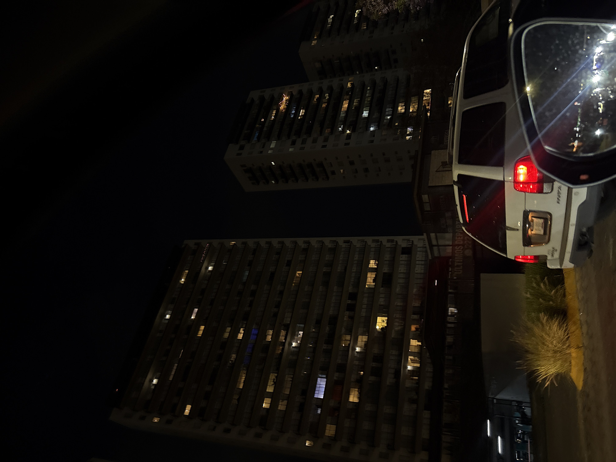 This image depicts a nighttime urban scene in Heroica Puebla de Zaragoza, Mexico. In the mid-right foreground, the rear of a white SUV is visible, its red brake lights illuminated. The vehicle's license plate is present but unreadable. The bottom right corner shows a blurred reflection of bright lights within the side mirror of the observing vehicle. A small cluster of light-colored ornamental grasses rests on a yellow-painted concrete strip in the mid-ground. The background is dominated by three tall, multi-story residential buildings set against a dark, clear night sky. The leftmost and most prominent building features numerous windows, many illuminated with warm yellow light, while some emit a cooler blue light. Balconies are visible along its facade. A partially visible sign beneath these structures reads "ULTIMOS DEPARTAMENTOS" and the number "22". No individuals are discernible within the frame.