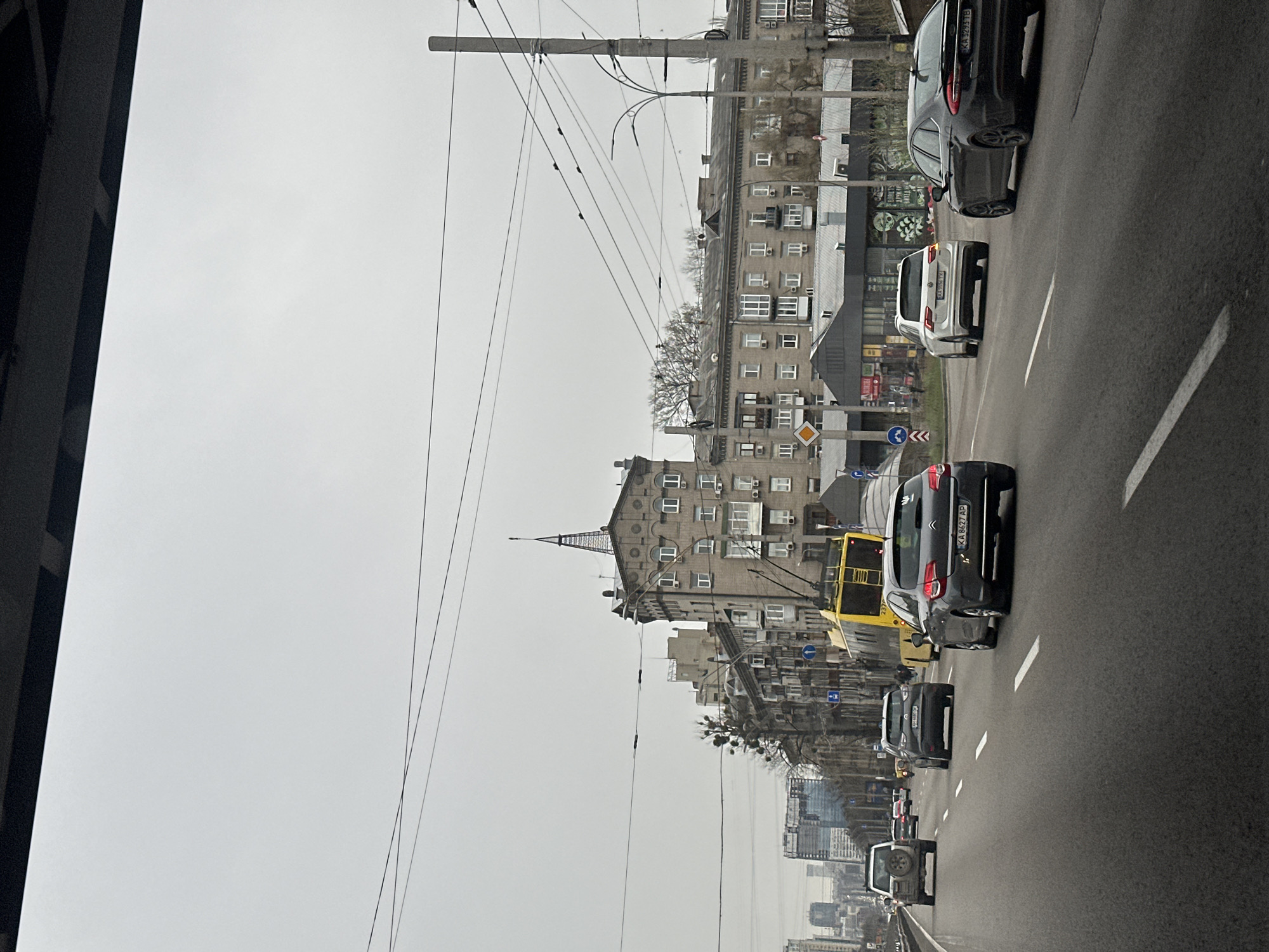 The image depicts a street scene in Kyiv, Ukraine on an overcast day. The view is captured from a high vantage point, likely an overpass or bridge, looking down onto a multi-lane road with vehicular traffic. Cars and a yellow trolleybus are moving along the road, adhering to marked lanes. On the roadside, there are Soviet-era buildings, identifiable by their architectural style and height. Overhead, a network of power lines and utility poles is present. In the distance, high-rise buildings can be discerned amidst a hazy urban landscape. The angle of the photo and composition suggests it was taken from a moving vehicle or pedestrian vantage point high above the street. The overall atmosphere is that of a typical urban weekday in Kyiv.