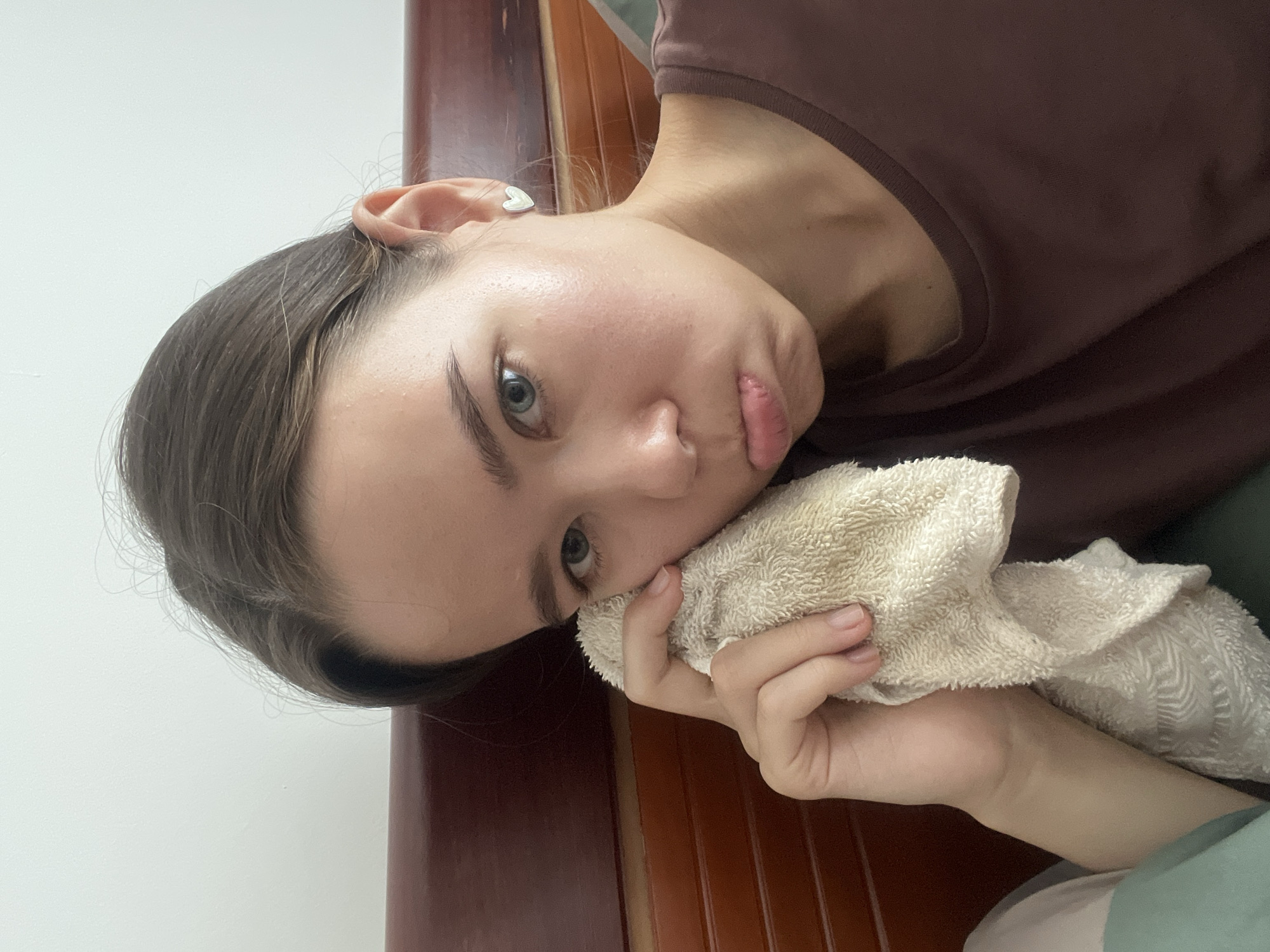 A young woman with brown hair and fair skin is seen from a close-up, angled perspective. She is holding a crumpled white towel to her face, with her left cheek resting on it. Her lips are slightly pursed. She's wearing a brown top. A white wall is visible in the background to the left, while a dark brown wooden surface appears behind her head and shoulder on the right. She has a heart-shaped earring in her ear. The environment does not appear to be in the city of Nam Natural Heritage Area Trang, Vietnam.