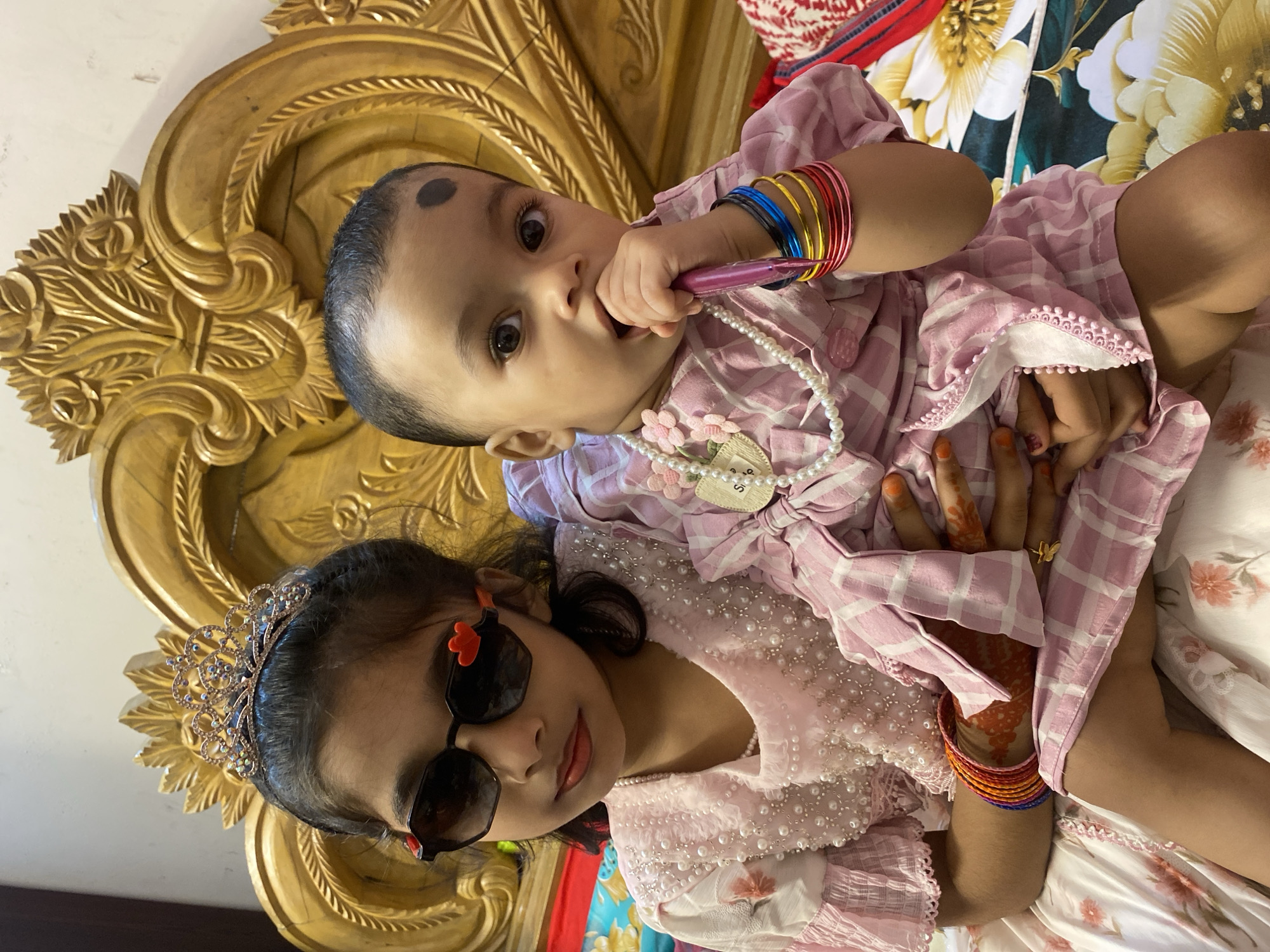 In a close-up, indoor shot taken in Dhaka, Bangladesh, two young girls are depicted. The girl on the left, older of the two, wears a tiara and dark sunglasses with red heart accents. She is adorned with a pearl necklace and a pink dress embellished with pearls. The younger girl, cradled by the older girl, is dressed in a pink and white checkered outfit and also wears a pearl necklace. She has a dark circular mark on her forehead and is holding a pink object to her mouth. Both girls have bangles on their wrists. They are seated on a bed with a decorative wooden headboard and floral patterned bedding. The lighting appears to be natural or artificial indoor lighting.