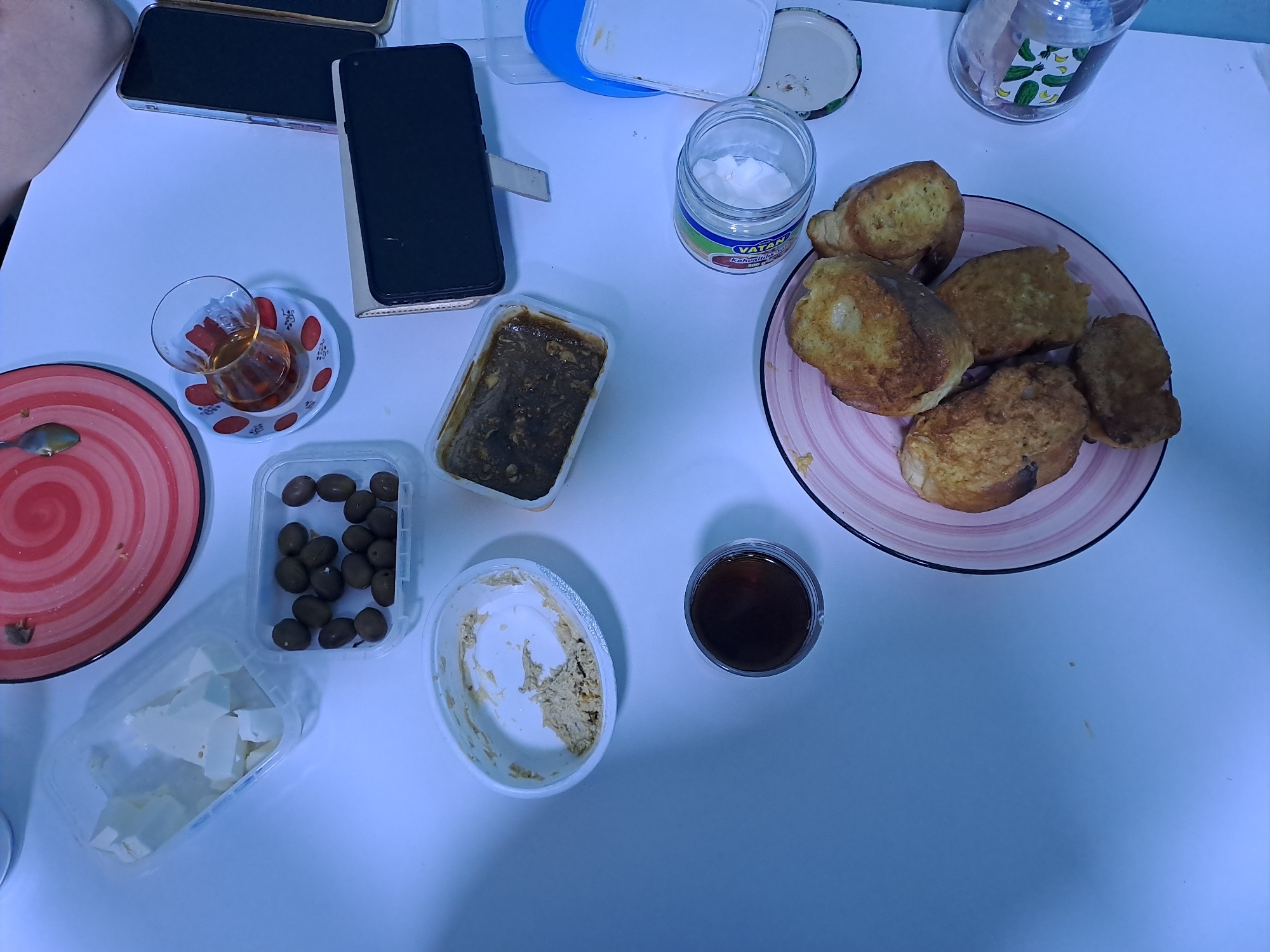 The image displays a breakfast spread on a white table. Several food items are visible, including a plate of fried bread or French toast, a container of olives, a dish with a dark, possibly savory, spread, a small glass of tea, a container of white cheese cubes, and a small bowl with a light-colored spread. A jar of what appears to be yogurt or cream cheese is also present, with a label that reads "VATAN Kahvaltilık". A second phone is lying on the table. The scene is set indoors, likely in a home, with bright overhead lighting. The time of day is not explicitly indicated, but the presence of breakfast items suggests it is morning. There are no obvious weather cues or strong emotional indicators. The overall impression is one of a casual breakfast setting.