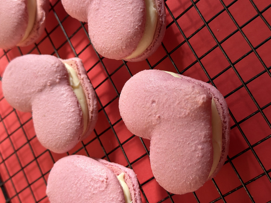The image displays six heart-shaped pink macarons with a pale yellow cream filling, arranged on a black wire cooling rack. The macarons are a light pink hue with a slightly textured, matte surface. The cooling rack rests on a solid, vibrant red background. No people, actions, or interactions are present in the scene. The environment appears to be an indoor setting. The specific location in Tel-Aviv, Israel, cannot be determined from the visual information provided.