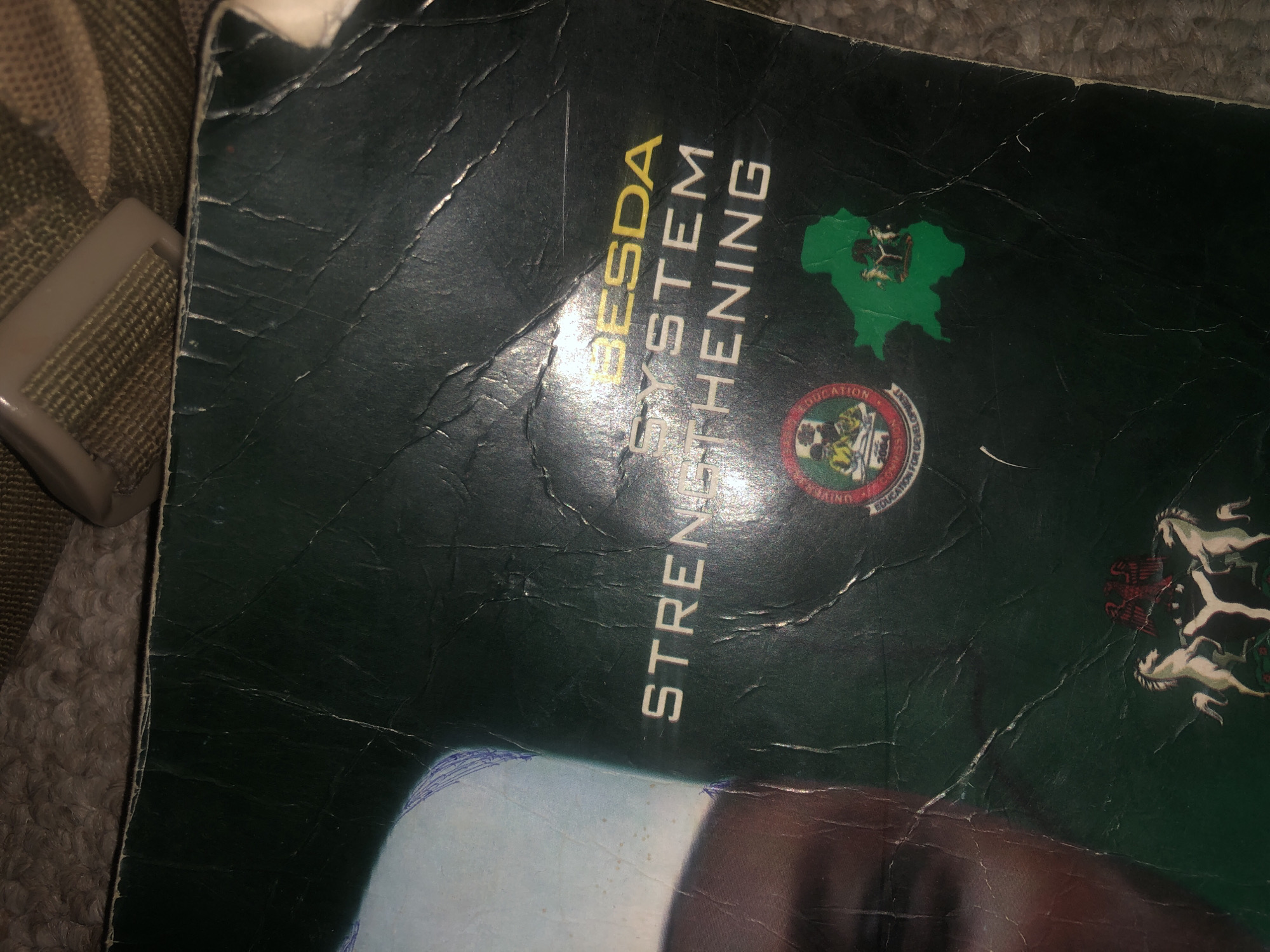 The image shows a close-up of a dark green book cover, possibly a report or publication. The cover is creased and worn, indicating age or heavy use. Dominant text on the cover reads "SESDA SYSTEM STRENGTHENING" in white and yellow lettering. Below this text, a circular emblem and a green map of a Nigerian state are visible. The emblem features figures and text that are difficult to decipher clearly due to the image quality. The green map is overlaid with another emblem, possibly a coat of arms, which includes two white horses, an eagle, and a shield. The background appears to be a textured surface, possibly carpet or fabric, in a neutral tone. Part of a person's face, with a light-skinned complexion, is visible on the left side of the image, obscured by shadows and the book cover. The overall impression is that of a document being photographed in a casual setting, possibly indoors. The specific location in Jalingo, Nigeria, cannot be determined from the visual information provided in the image.