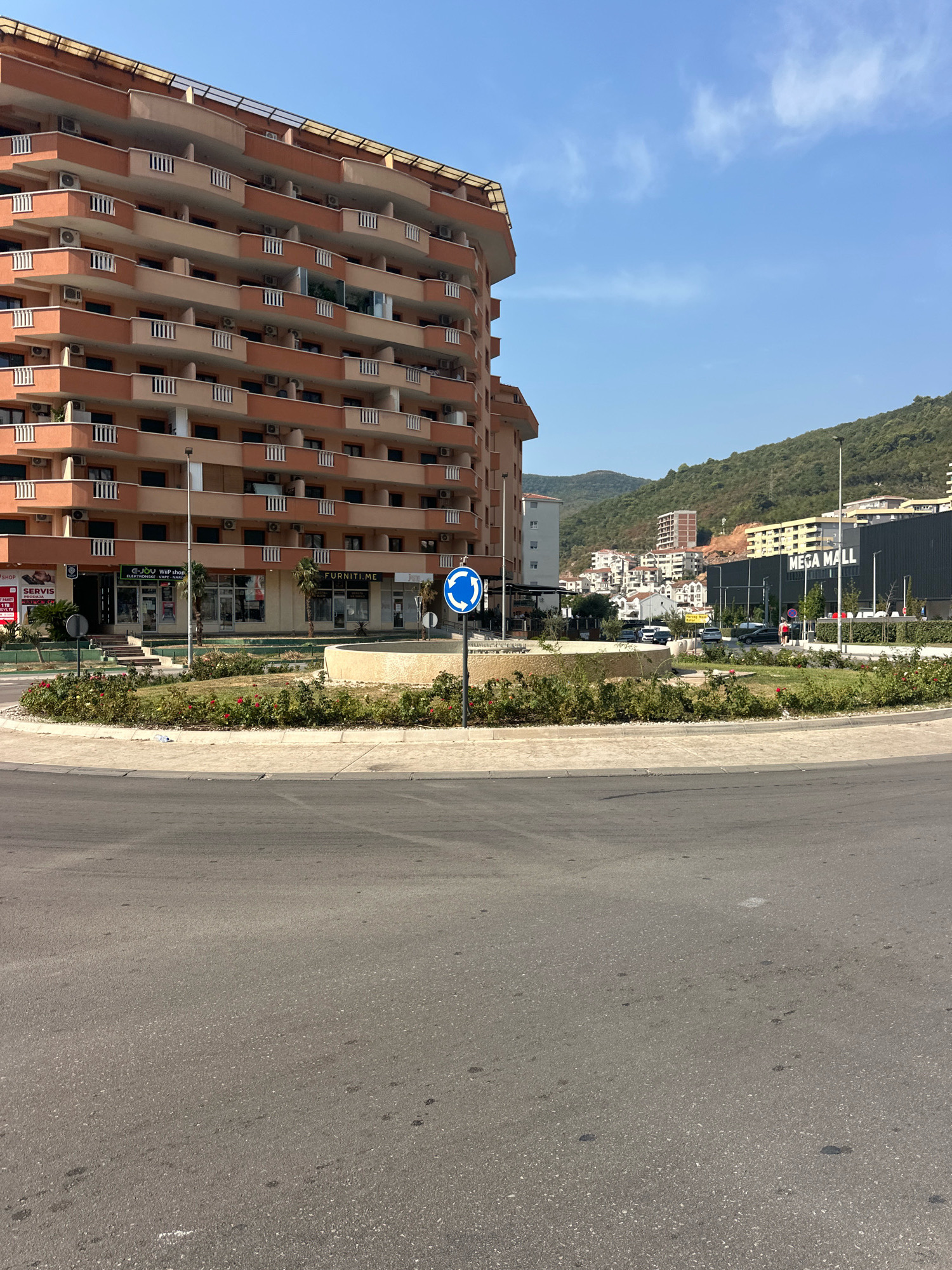 The image shows a view of a roundabout in Budva, Montenegro. On the left is a large, multi-story building with many balconies. In the center of the roundabout is a circular garden with red roses and other plants, and a low, beige fountain structure. A blue traffic sign indicating a roundabout is visible. Across the roundabout, a large, modern building with "MEGA MALL" written on its facade is prominent. Behind the mall, a hillside is covered with buildings and lush green vegetation. The sky is clear and blue. The foreground is dominated by asphalt road.