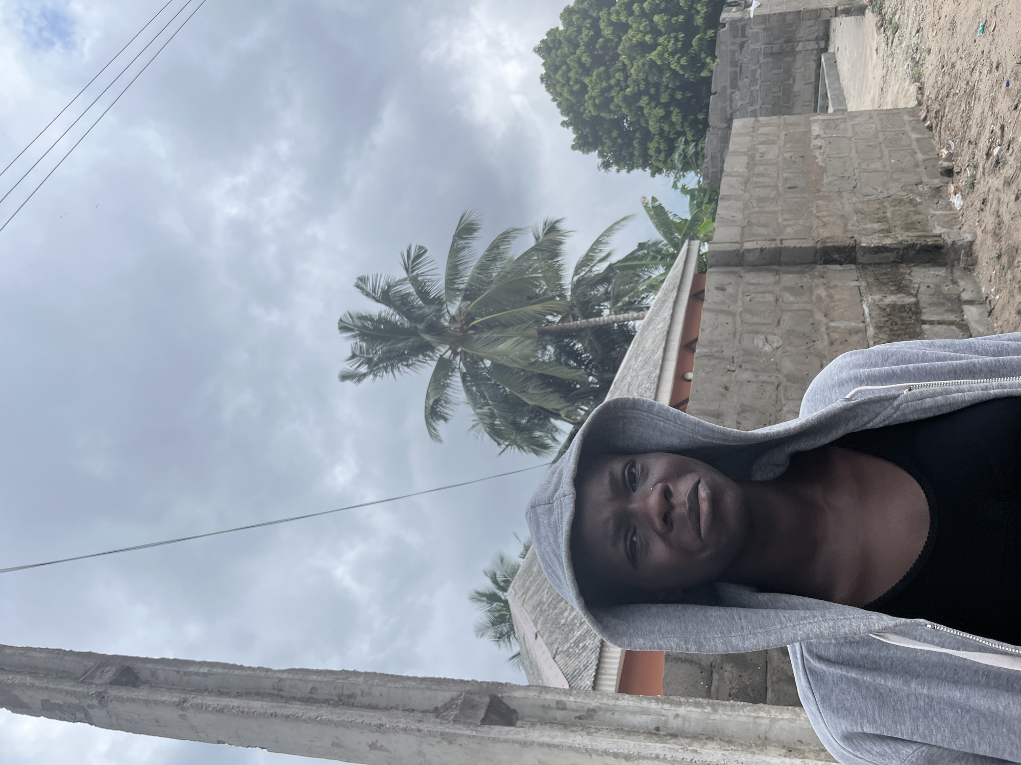 A medium shot, taken at an extreme angle, captures a person wearing a gray hooded sweatshirt, gazing directly at the camera. The individual is wearing a black top underneath the sweatshirt. The setting appears to be outdoors, with a partially constructed concrete building visible. A tall palm tree is centered in the background, partially obscured by the clouds. Overhead electrical wires stretch across the cloudy sky. Lush green trees and shrubs appear in the upper right corner. The image appears to be taken in Badagry, Nigeria.