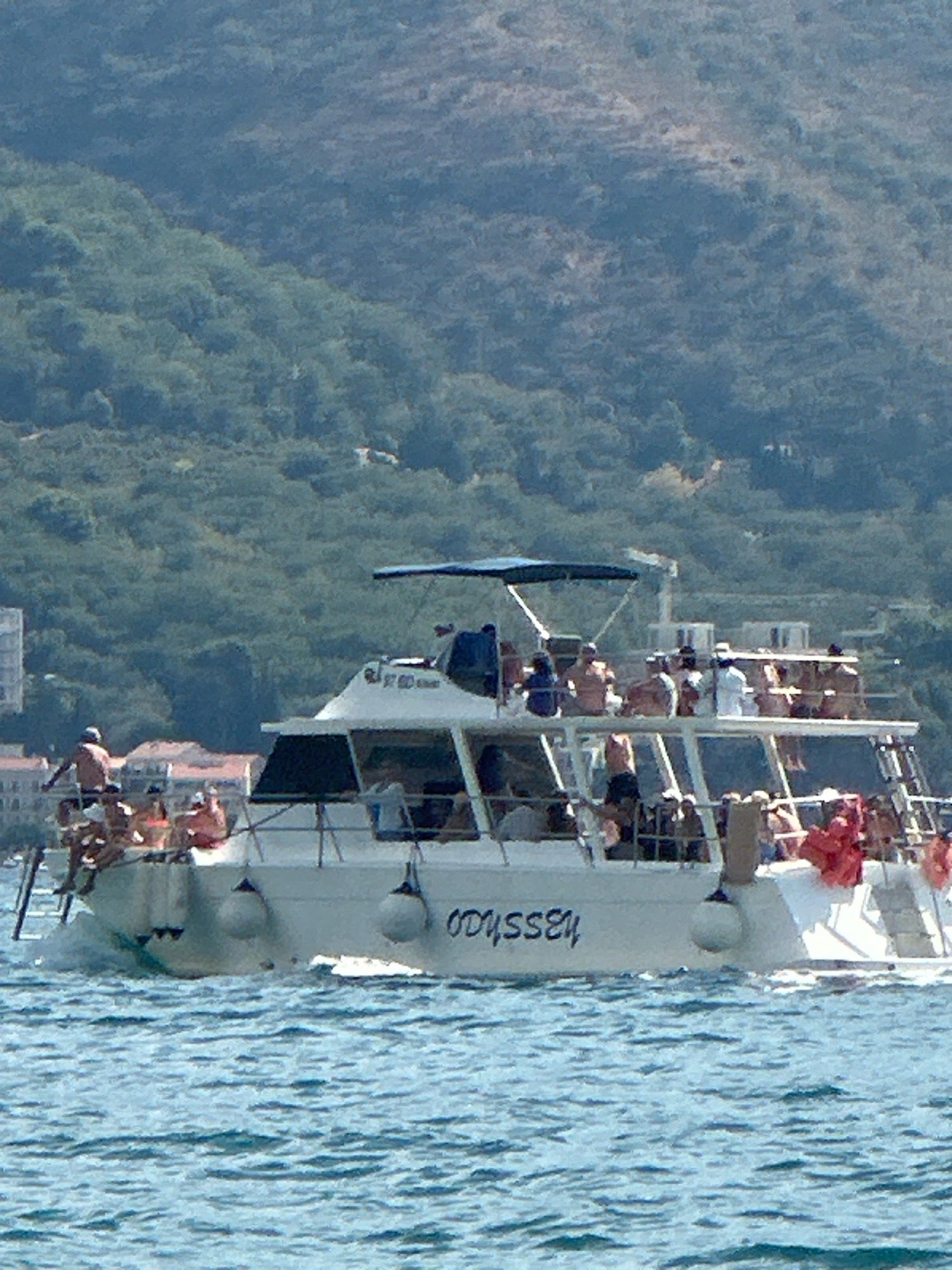A wide-angle, vertical shot captures a white, multi-level passenger boat named "ODYSSEY" sailing on a bright blue, wavy sea in front of a lush, green, mountainous backdrop. The boat appears to be full of people, many of whom are visible on the upper decks, some standing and others sitting. The people are dressed casually, and some appear to be wearing swimwear. There are life preservers hanging on the side of the boat, and a ladder is visible on the left side. The boat is creating a wake as it moves through the water. In the background, the green hills are dense with foliage, and some buildings are visible on the coastline below the hills, suggesting an urban or semi-urban setting. The sky is a clear, light blue. The overall scene depicts a leisurely day on the water, likely a tourist excursion.