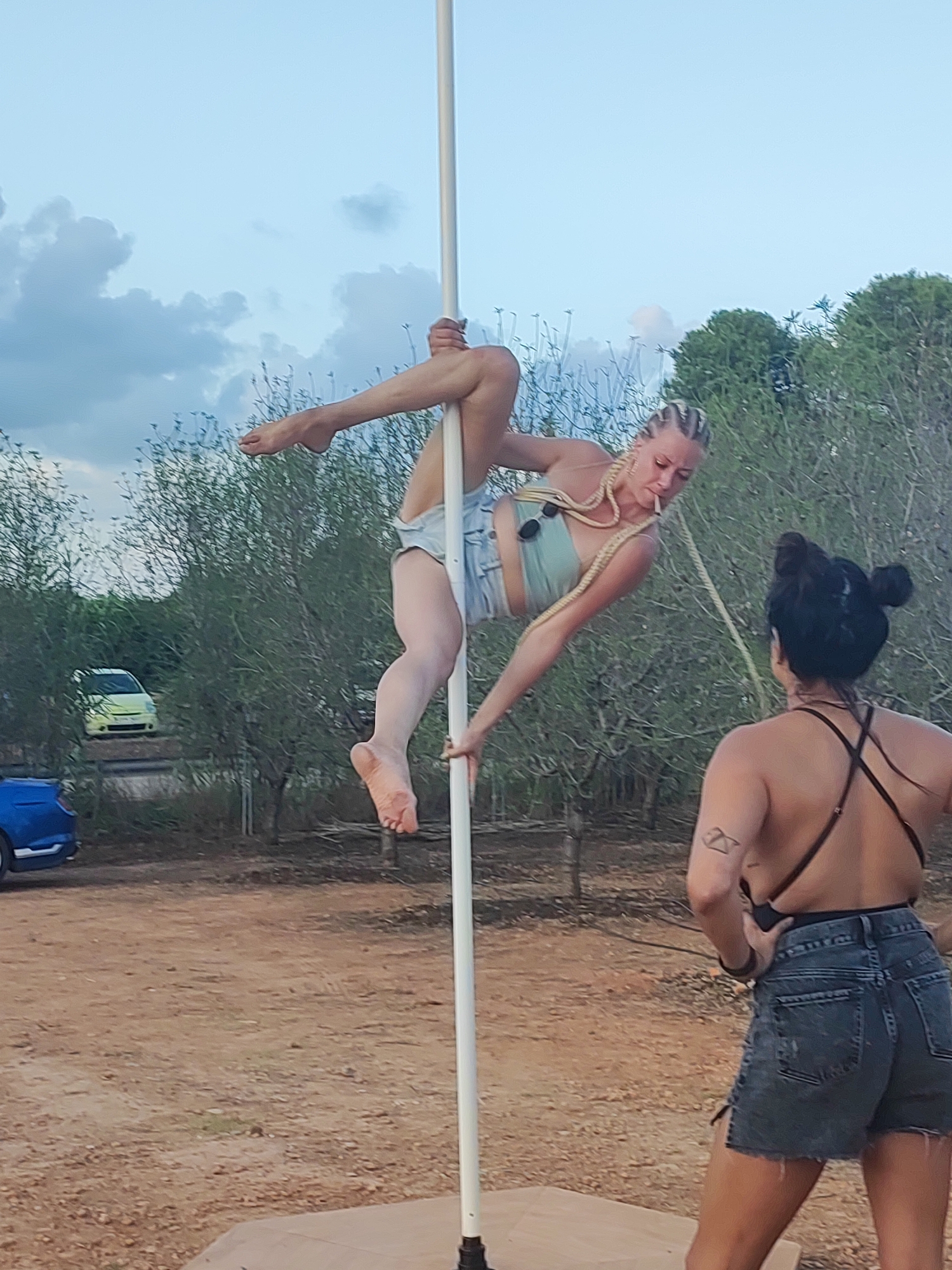 In an outdoor setting, two women are visible. The woman on the left is performing a pole dance routine, suspended in mid-air by a vertical pole. She has blonde hair styled in braids and is wearing a light blue crop top and denim shorts. The woman on the right is facing away from the camera, observing the performance. She has dark hair styled in a bun and is wearing a dark-colored top with thin straps and denim shorts. The environment appears to be a rural area with dry, dusty ground and sparse vegetation, including trees. Two vehicles, a light yellow car and a blue car, are parked in the background. The sky is partly cloudy, suggesting it is either early morning or late afternoon. The location is identified as Alcàsser, Spain.