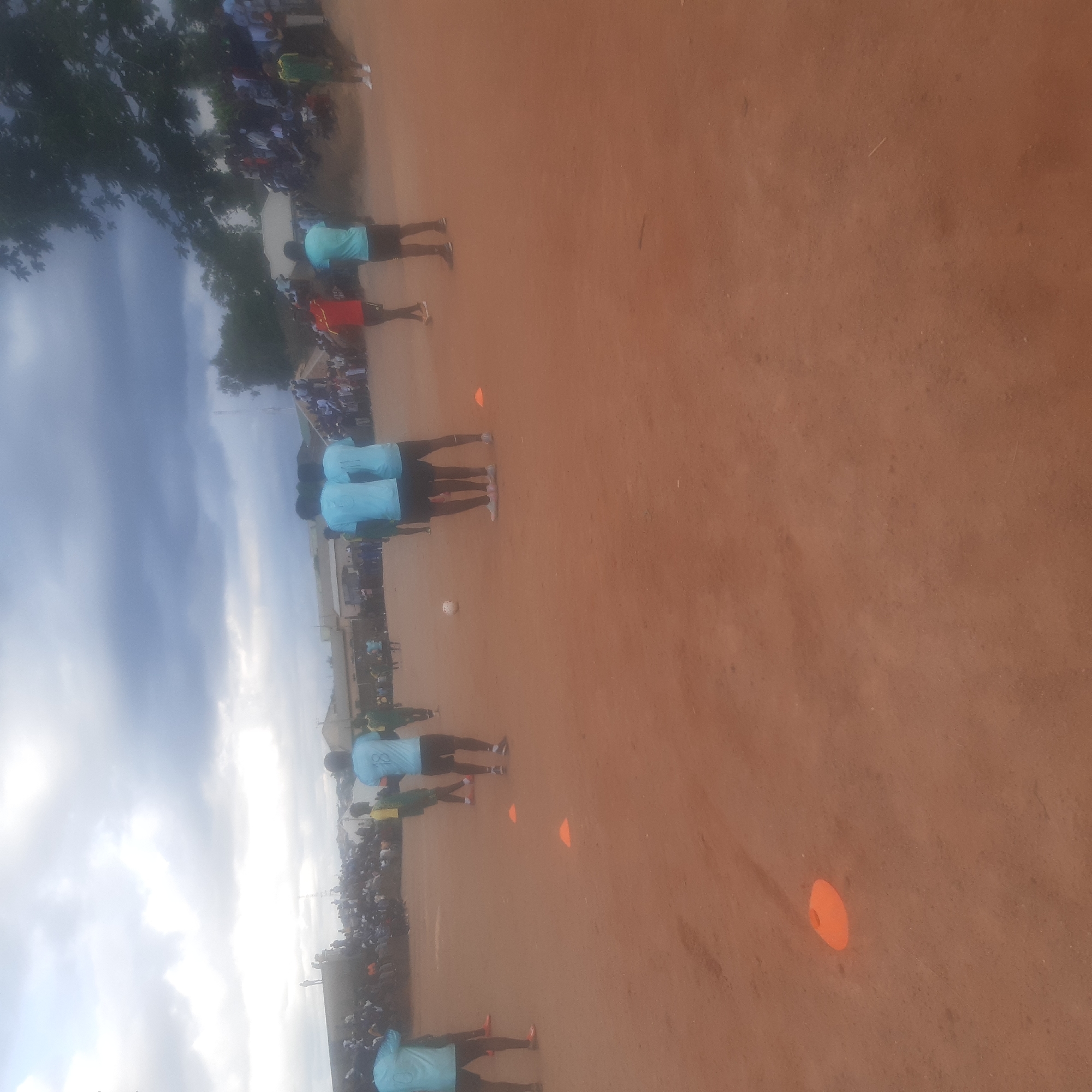 The image depicts an outdoor scene on a dusty, reddish-brown ground, likely a football pitch, under a cloudy sky. In the foreground, several orange cones are scattered, suggesting training or practice drills. A white soccer ball is visible in the middle ground.
A group of young men, presumably athletes, are present. Several are wearing light blue jerseys with numbers on the back, and dark shorts. Others are in green jerseys. Some are positioned around the ball, while others are further back. Spectators are visible in the background, gathered on a raised embankment and behind a fence, indicating a public event or a community gathering. The environment appears to be an open field, possibly in a less developed urban area, with low-rise buildings and trees visible in the distance. The overall impression is of a football match or training session taking place in Jos, Nigeria.