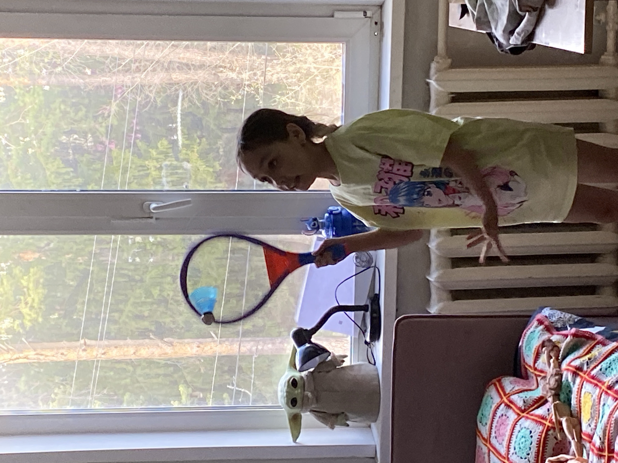 This image captures a young person indoors in Votkinsk, Russia, engaged in a playful activity near a window. The subject, a child or teenager with dark hair, is wearing a light yellow-green t-shirt featuring an anime-style graphic with Japanese text, including visible characters like "田" and "桜". They are holding a purple and red badminton racket, with a blue shuttlecock positioned on or near the strings, as if caught or about to be hit. The person's expression is one of focused engagement.
The scene is brightly lit by natural daylight streaming through a large window, suggesting it is daytime. Outside the window, a green, treed landscape with some bare branches is visible, possibly indicating late winter or early spring foliage. On the white windowsill, a "Baby Yoda" (Grogu) figurine is prominently displayed, holding a small, black adjustable desk lamp. A blue plastic object, possibly a toy or bottle, is also on the windowsill.
To the right of the person, a white, sectioned radiator is situated below what appears to be a bed or couch, partially covered by a vibrant, crocheted blanket with a multi-colored geometric pattern (squares in white, green, pink, red, and yellow). A wooden articulated human figure rests on this blanket. Some grey fabric or clothing is draped over the top of the radiator. The overall impression is a cozy, lived-in room.