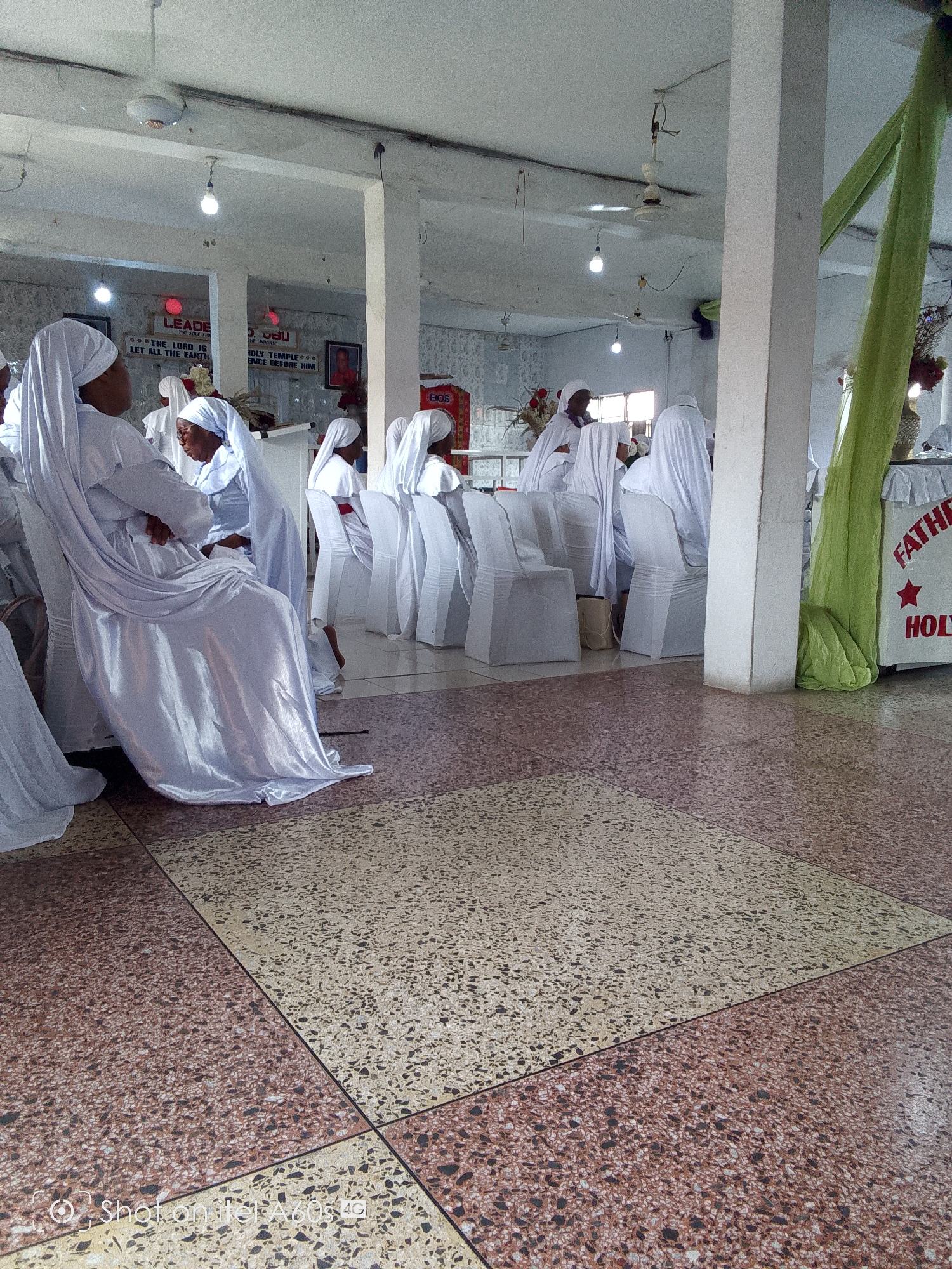 A wide-angle shot depicts an interior setting with several individuals dressed in white attire, presumably attending a religious gathering. The location appears to be a large hall, characterized by tiled flooring and white-painted structural columns supporting the ceiling. The individuals, seated in white-covered chairs, are positioned in rows, with some engaged in conversation. A sign with the words "FATHER" and "HOLY" is visible. There are decorations. The hall has a ceiling fan, and lights hang from the ceiling. The photo was taken on an Itel A60s. The location appears to be in a city named Unknown, Unknown.