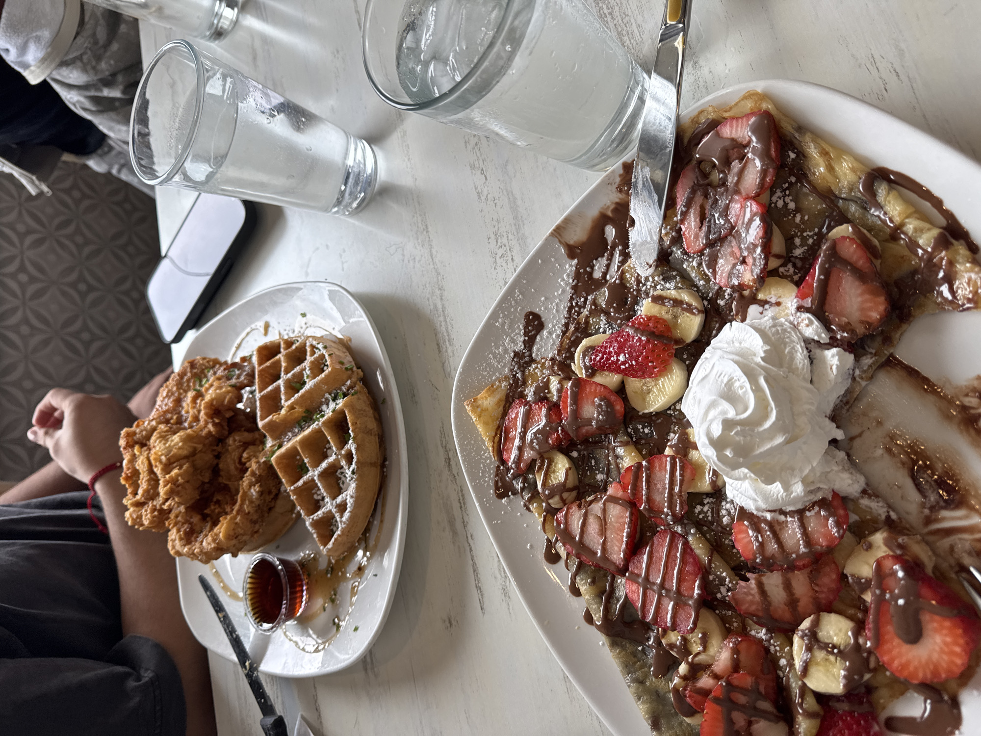 The image shows a table with food and drinks. On the left is a plate with fried chicken and a waffle, accompanied by a small container of syrup. To the right is a crepe topped with strawberries, bananas, chocolate sauce, and whipped cream. There are also three glasses of water on the table. The location is in St. Charles, United States of America. There is a person seated at the table, visible by an arm and hand.