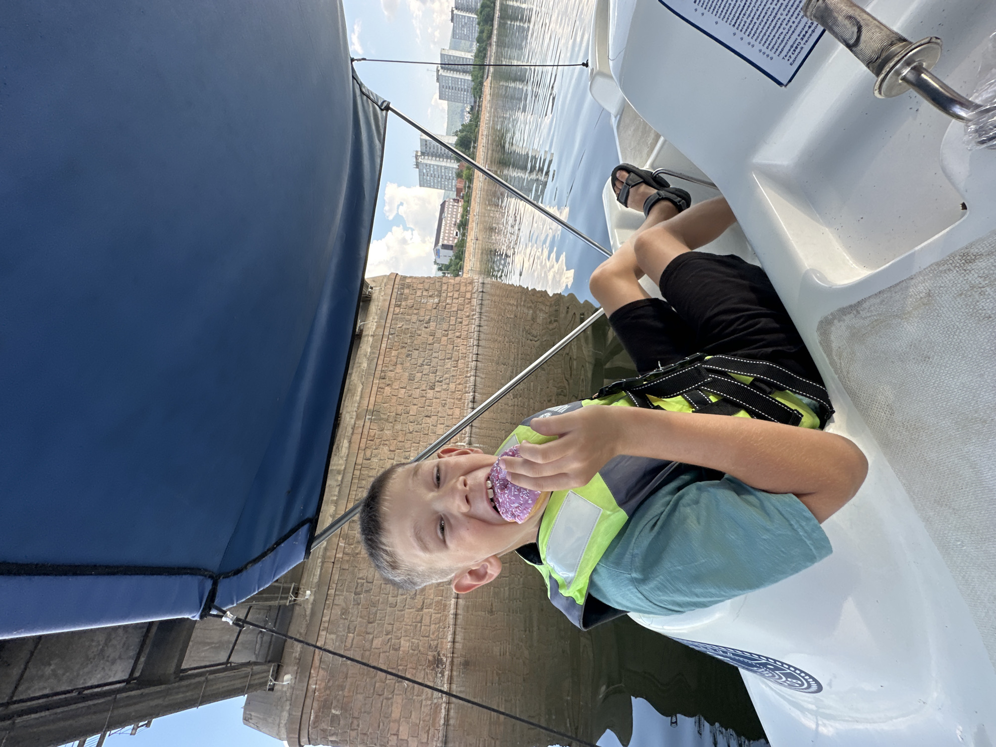 A young boy, wearing a green t-shirt and a life vest, is seated in a white boat on a body of water. He is holding a purple donut to his mouth and appears to be taking a bite. The boat has a blue canopy overhead. In the background, a large stone bridge and several buildings are visible on the shore. The water reflects the sky and the surrounding structures. The boy is wearing black shorts and sandals.