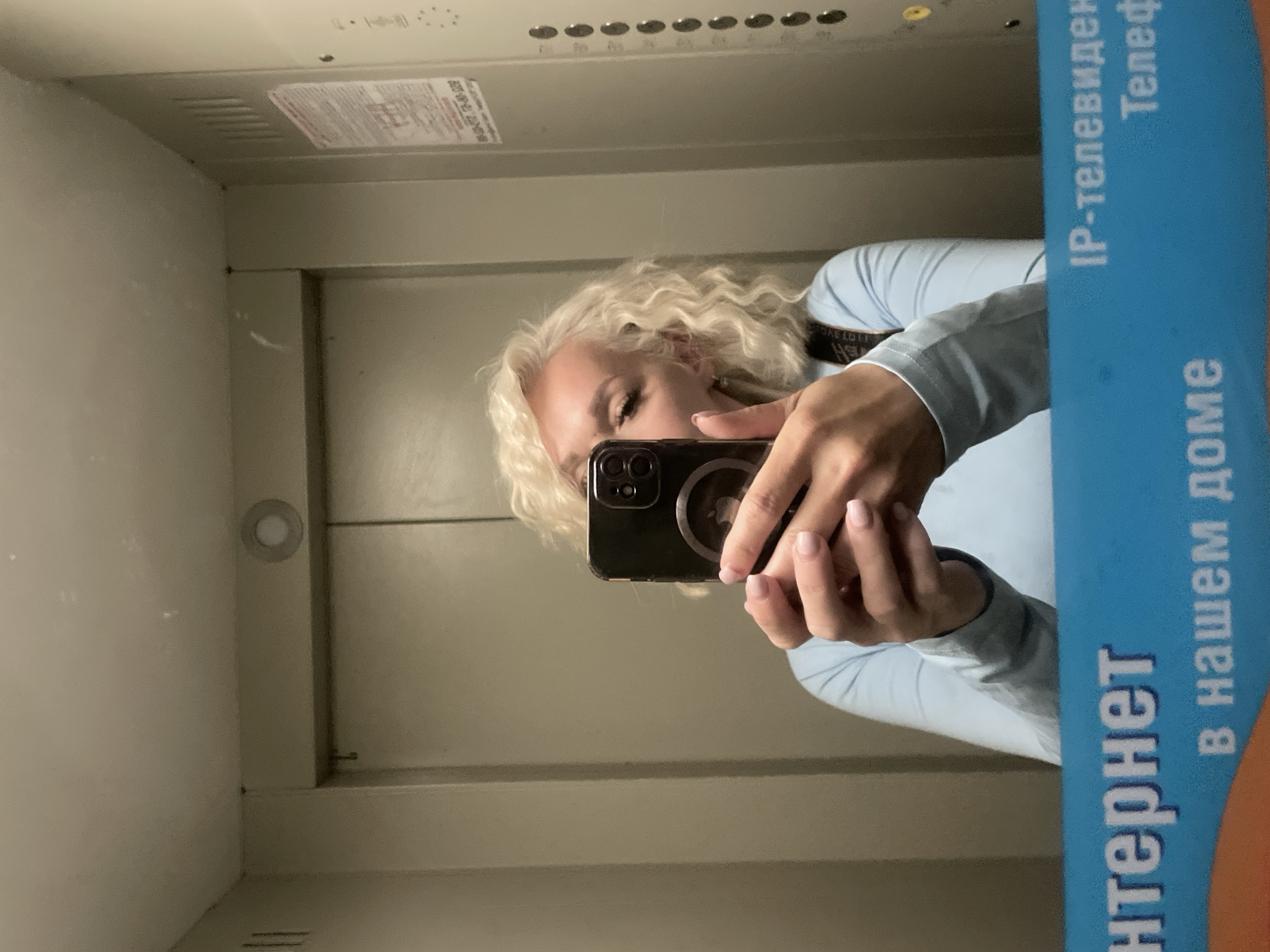 A woman with blonde curly hair is taking a selfie in an elevator. She is wearing a light blue long-sleeved shirt. She is holding a black smartphone in her hands, which is reflecting in the mirror. The elevator has metallic doors and beige walls. There is a control panel with buttons on the right side of the elevator. A blue banner with text in Russian is visible at the bottom of the image.