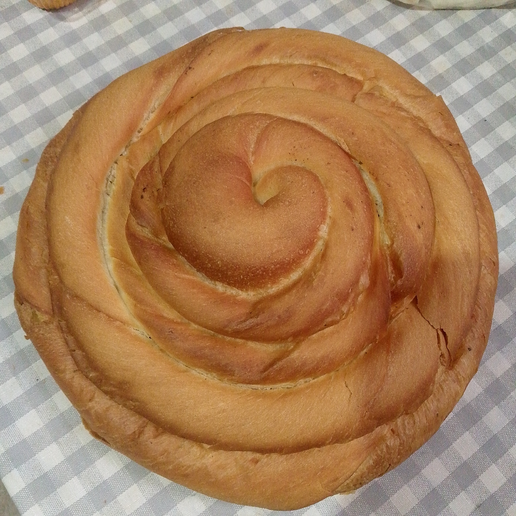 A close-up, overhead shot captures a golden-brown, spiral-shaped bread loaf placed on a white and light blue checkered tablecloth. The bread's crust is slightly darker in some areas, suggesting it is baked through. The light gray and white checkered pattern of the tablecloth is visible in the background. There are no people or other objects present in the scene. The image appears to be taken in an indoor setting, possibly a kitchen or dining area.