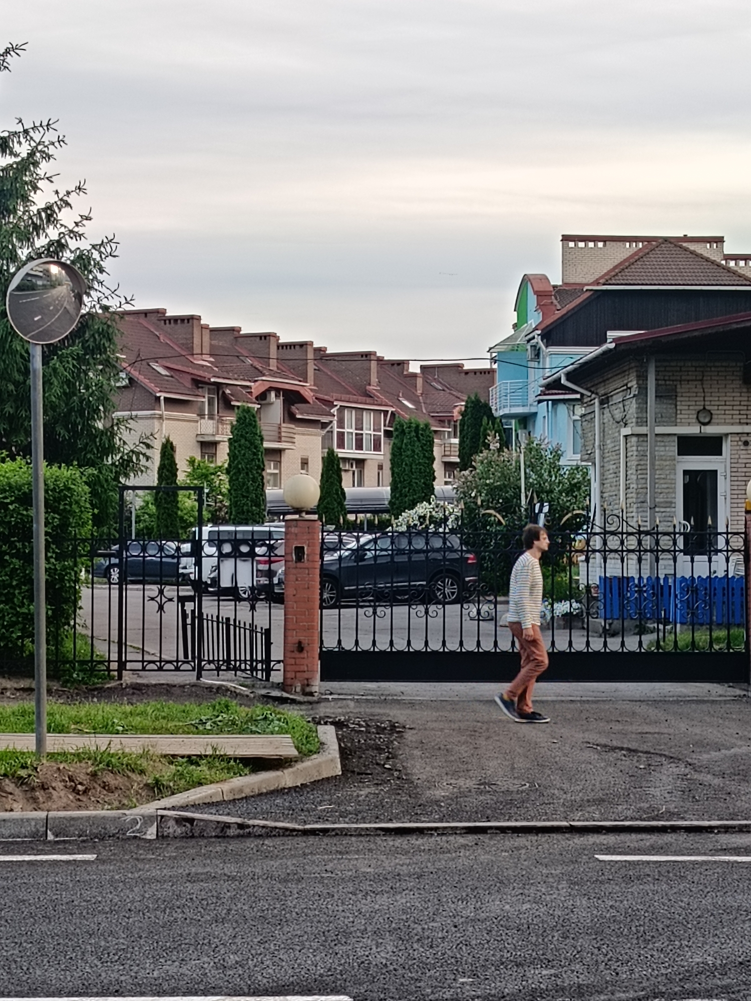 This image depicts a residential street scene in Saint Petersburg, Russia, under an overcast sky.
In the foreground, an asphalt road runs horizontally, featuring a visible dashed white lane marking. A concrete curb separates the road from a narrow, unkempt grassy verge with exposed soil and a wooden plank lying partially across it. On the far left, a grey metal pole supports a circular convex traffic mirror reflecting a section of the road.
A single adult individual is walking from left to right across the asphalt. The person wears a light-colored horizontally striped long-sleeved shirt, reddish-brown trousers, and dark athletic shoes. Their left foot is lifted mid-stride, and their head is turned slightly left with a forward gaze.
Beyond the road and verge, a tall, black, ornate wrought-iron gate with decorative four-pointed star motifs spans the midground. A smaller pedestrian gate within the main gate is open on the left. A brick gate post, topped with a spherical light fixture, stands to the right of the pedestrian gate.
Behind the gate, a paved driveway area contains several parked automobiles, including a white SUV and multiple dark-colored sedans or SUVs. Various vegetation is present, including a dense evergreen bush on the far left and several tall, slender coniferous trees (possibly cypresses or junipers) arranged in rows behind the gate, interspersed with other bushes and flowering plants with white blossoms.
In the background, a row of multi-story residential buildings with brown tiled pitched roofs and numerous chimneys is visible. These structures feature light-colored brick facades, some with balconies and dormer windows. To the right, a distinct light blue house with white trim and multiple balconies stands adjacent to a grey brick building with a dark-colored roof section in the closer background. The sky is uniformly light grey and overcast, indicating a cloudy atmospheric condition.