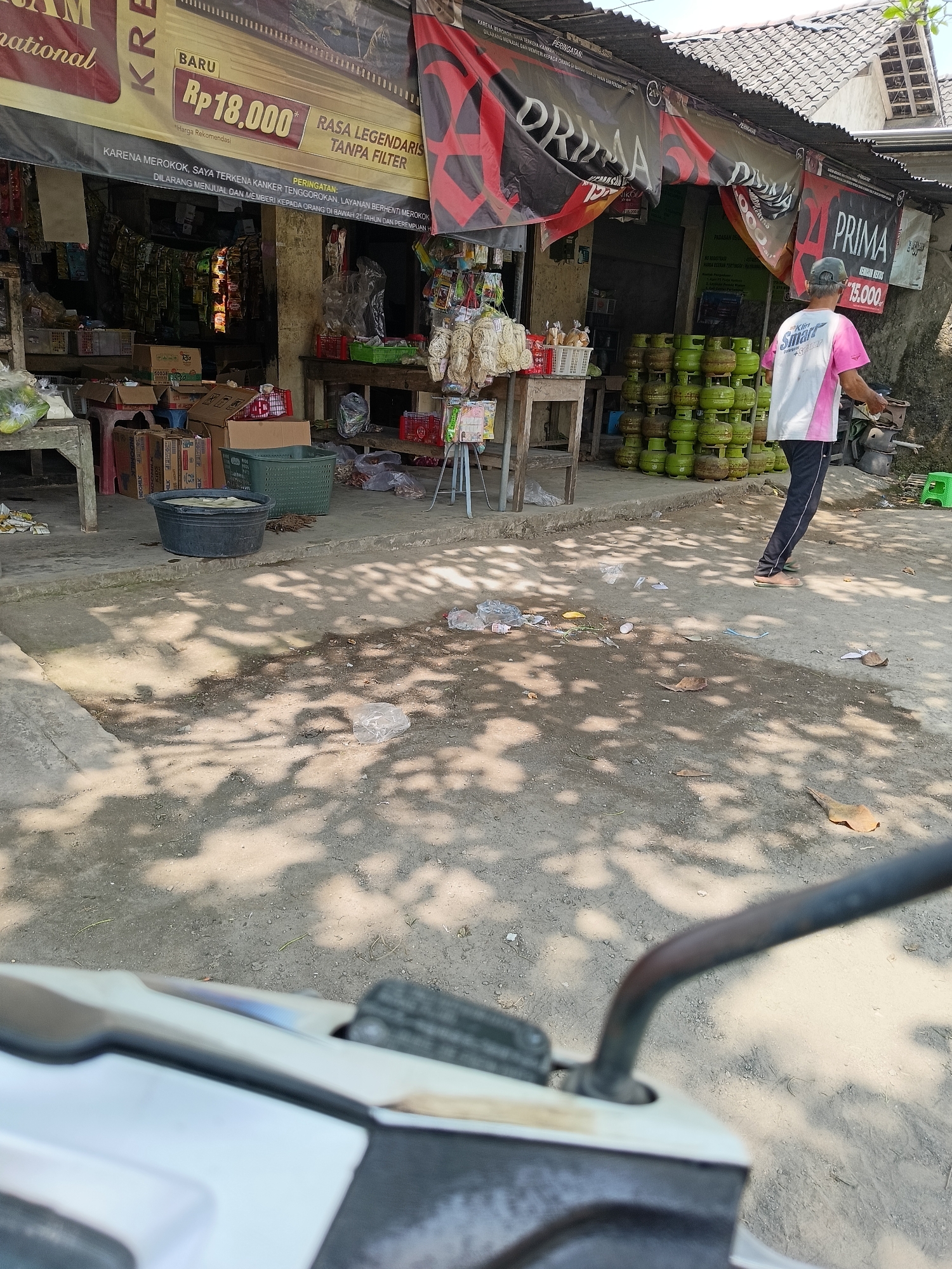 The image depicts a street-level view of a small shop in Barukan, Indonesia. The shop appears to be a convenience store or a local market, selling various goods. In the foreground, a motorcycle's handlebars are visible, indicating the photographer's perspective. The ground is dirt and gravel, with scattered debris like leaves and plastic.
The shop itself is covered by a corrugated metal roof and has an awning displaying banners. One banner prominently features the word "PRIMAGROUP" with the price "RP 18.000" displayed on a yellow background, suggesting a promotion. Another banner states "PRIMAGROUP" again, with "Rp 15.000" below it. Red and black banners are also present, one with a large "X" symbol.
Inside the shop, shelves are stocked with packaged goods, including what appear to be snacks and possibly cigarette packs. Green gas cylinders are stacked neatly outside the shop, indicating they are also for sale.
A person, likely a man wearing a pink and white shirt with "Smart" written on the back and dark pants, is walking away from the shop. He is wearing sandals. The environment is outdoors, with sunlight casting shadows on the ground, suggesting it is daytime. The overall impression is that of a typical small business operating in a local Indonesian community.