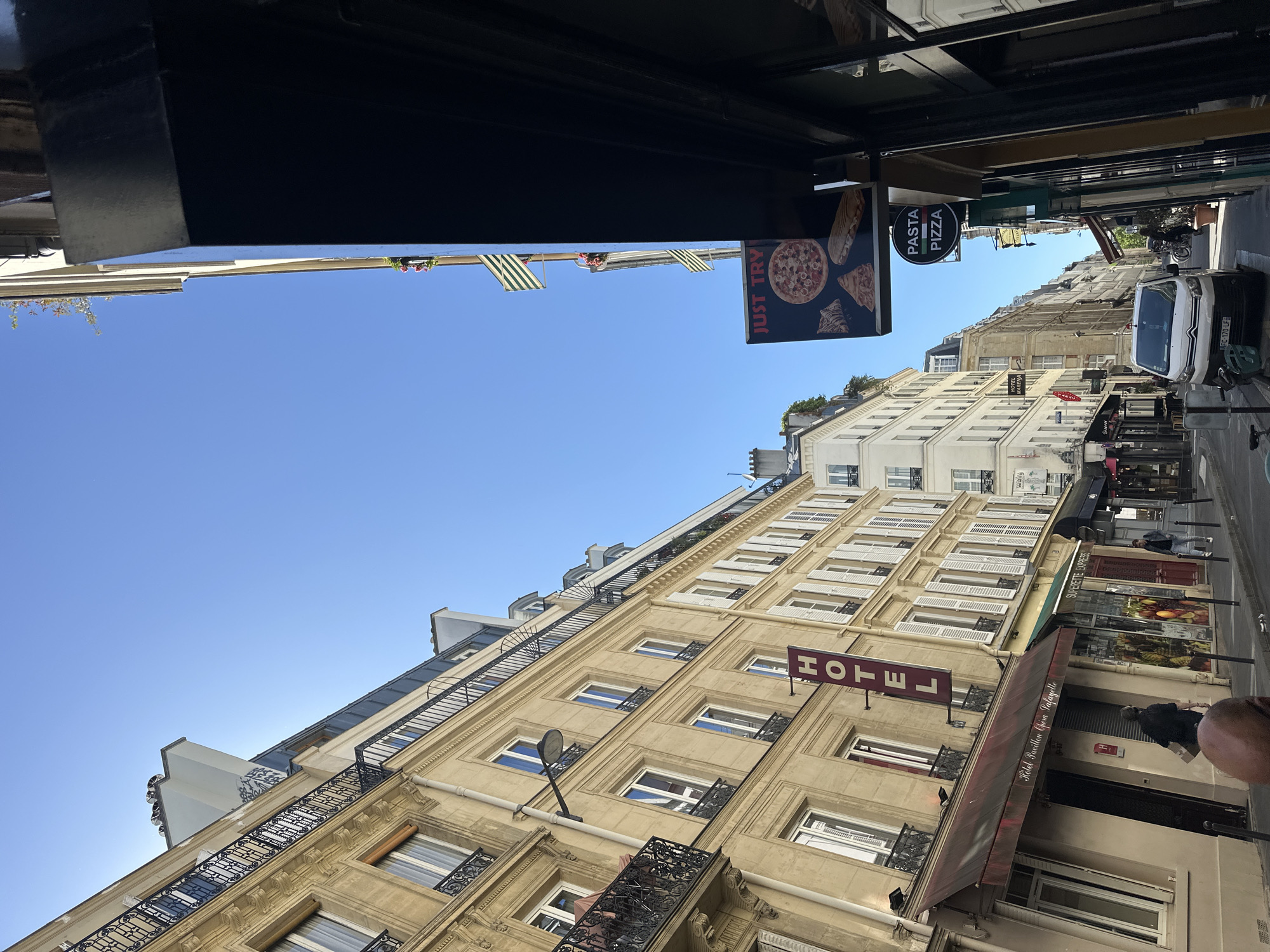 This media file captures a bright, sunny daytime scene on a narrow street in Paris, France. The perspective is looking down a street lined with multi-story, classic Parisian buildings, characterized by their light-colored facades, numerous windows, and wrought-iron balconies. The sky above is a clear, vibrant blue.
On the left side of the street, a prominent hotel building features a large, vertical red sign displaying "HOTEL". Below this, another sign reads "Hotel Pavillon Opéra Lafayette", indicating its specific location. Next to the hotel entrance is a smaller sign with an "H" logo. Further down, there's an establishment named "SUPERETTE EXPRESS" with a display of fruits and vegetables.
On the right side, another building features awnings and a sign for a food establishment. A black hanging sign advertises "JUST TRY" and "PASTA PIZZA", accompanied by images of pizza slices and a baguette. The street itself contains a parked white car with the license plate "FC-170-LF", and a scooter or motorcycle can be seen further down. A few pedestrians are visible, one person walking near the hotel entrance and another in the distance. The street appears calm, suggesting a typical daily urban scene. Overall, the image conveys a sense of everyday life in a European city, specifically Paris.