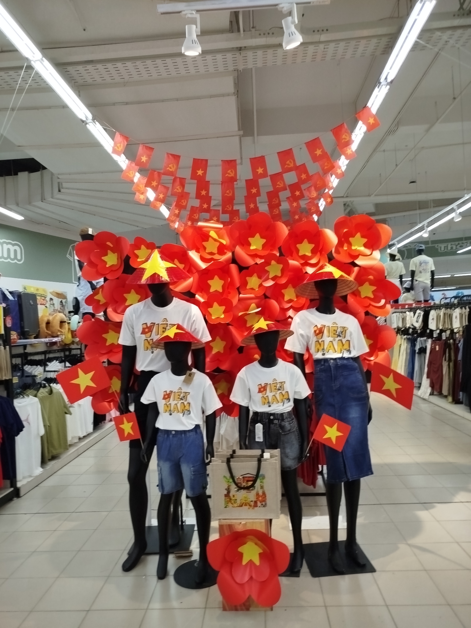 This image captures a patriotic merchandising display inside a retail store, likely a supermarket or department store, in Da Lat, Vietnam. The central focus is a cluster of red paper flowers, each with a yellow star at its center, arranged around four black mannequins.
Two adult and two child-sized mannequins are featured. All are wearing white t-shirts with "VIỆT NAM" printed in bold yellow letters. They are also adorned with traditional Vietnamese conical hats (nón lá), which have been decorated with a yellow star, resembling the Vietnamese flag. The adult female mannequin wears a blue denim midi skirt, holding a small Vietnamese flag. The adult male mannequin wears blue cargo shorts. The child mannequins wear dark denim shorts and blue denim shorts respectively, also holding small Vietnamese flags. A reusable tote bag with illustrations rests near the child mannequins.
Overhead, strings of small flags are suspended across the ceiling. These flags alternate between the red and yellow star flag of Vietnam and red flags featuring the yellow hammer and sickle symbol, representing the Communist Party of Vietnam. The ceiling features fluorescent strip lighting and white panels.
In the background, typical retail shelving and clothing racks display various garments. A partial "om" from a larger sign is visible on the left wall, suggesting a store name. The overall display emphasizes national pride and identity through the prominent use of Vietnamese symbols and colors. The artificial lighting indicates an indoor setting.
