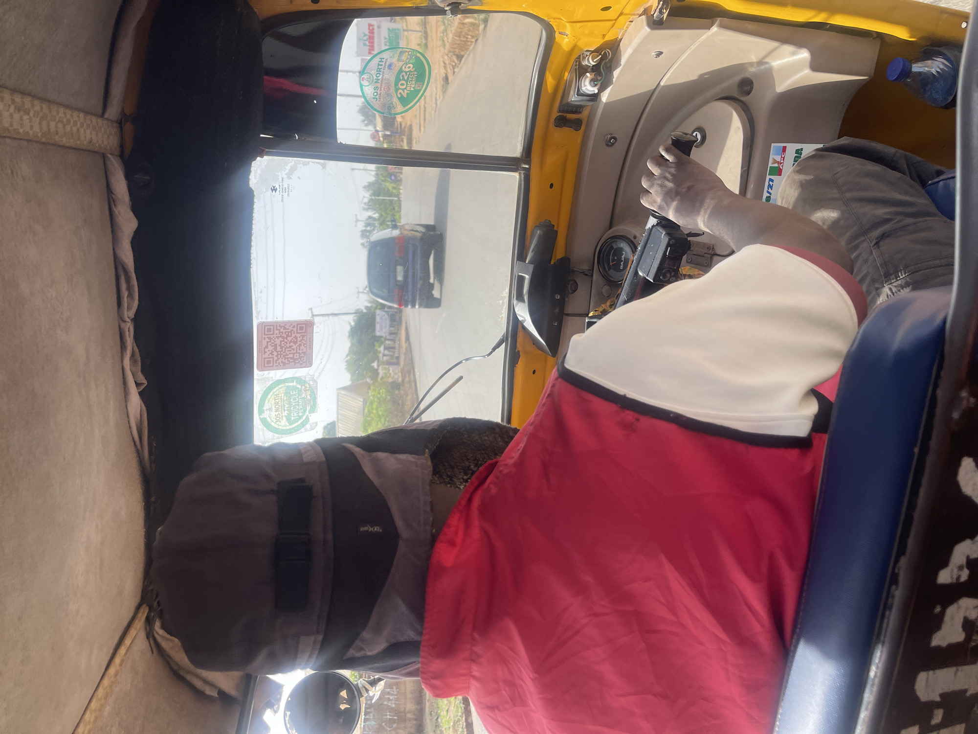 The image provides an interior perspective from a yellow auto-rickshaw, looking forward through the windshield. The driver is visible from behind, occupying the right side of the frame. They are wearing a white or cream short-sleeved shirt with red sleeves or trim, and grey trousers. Their left hand rests on the handlebar-style steering control. A dark grey bag or piece of clothing is positioned behind the driver's head, on the seat. The dashboard displays an instrument cluster with the "BAJAJ" brand name visible, and a plastic water bottle with a blue cap is situated near the driver's right shoulder.
Through the windshield, a paved road stretches ahead under a clear, bright sky, indicating daytime. A dark-colored car can be seen in the mid-distance on the road. Sparse vegetation and distant buildings line the roadside.
Several stickers are affixed to the windshield and dashboard:
* A prominent green circular sticker on the top left reads "JOS NORTH TRICYCLE PERMIT 2026," confirming the vehicle type and the location as Jos, Nigeria.
* A QR code sticker is visible below the permit sticker.
* A partial sign reading "FLY PHARMACY" is at the top right.
* On the dashboard near the driver, a small sticker shows the logo for "APC" (All Progressives Congress), a Nigerian political party.
The vehicle's interior features light beige or grey fabric lining on the ceiling and side panels, and a blue seat is visible to the driver's right. The scene depicts the vehicle on a road during daylight, likely in motion or briefly stopped.
