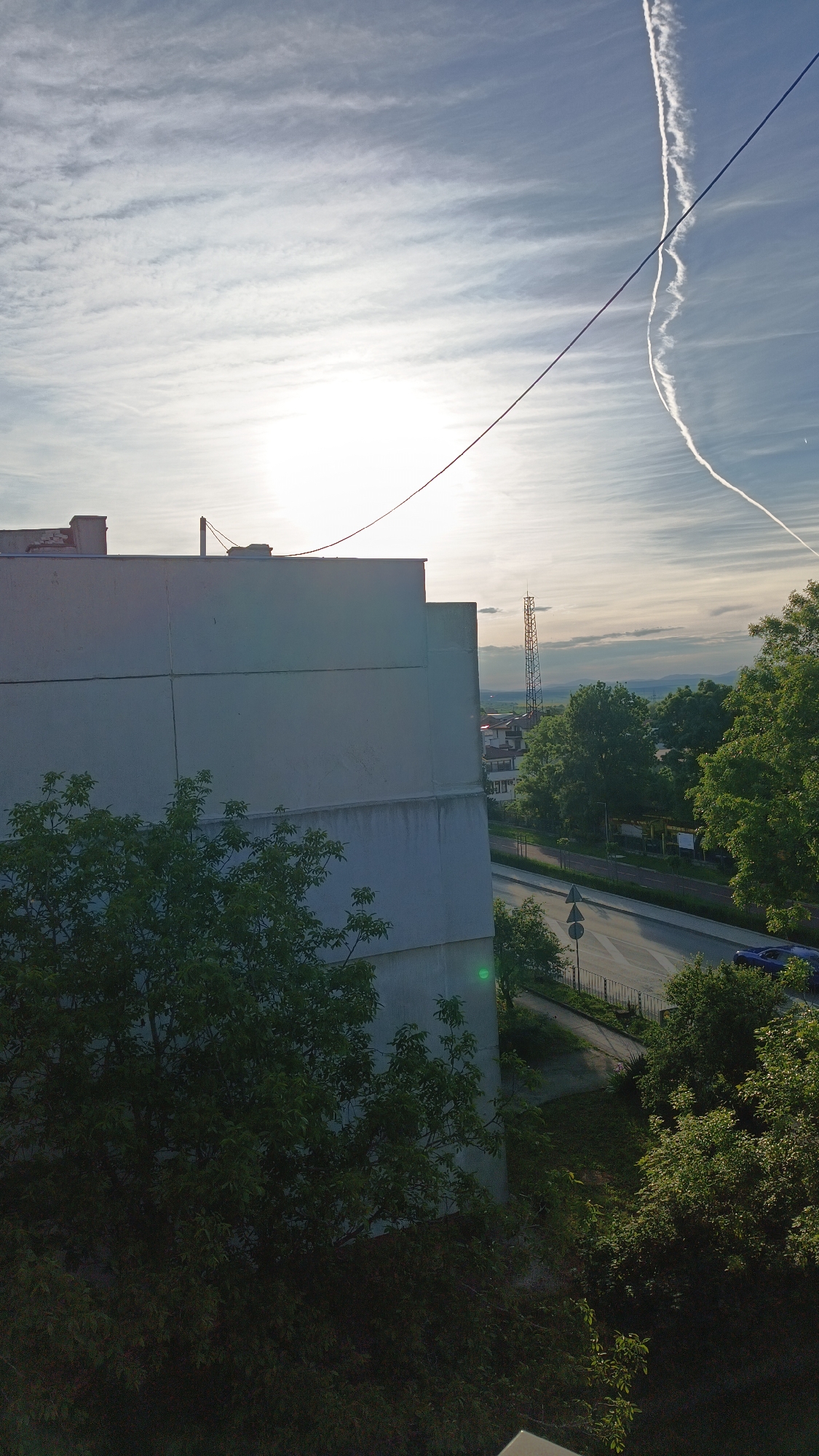 The image shows an outdoor scene from what appears to be an elevated position, possibly a window or balcony. A large, light-colored building dominates the left side of the frame, with its exterior showing some wear and discoloration. A tree with green foliage is partially obscuring the base of the building. To the right, a street runs alongside a sidewalk with trees lining the edge. A blue car is visible on the road. In the distance, a telecommunications tower is prominent, and beyond that, hills or mountains are visible under a bright sky with streaks of clouds and a contrail from a jet. The city of Karnobat, Bulgaria is not explicitly evident within the image.
