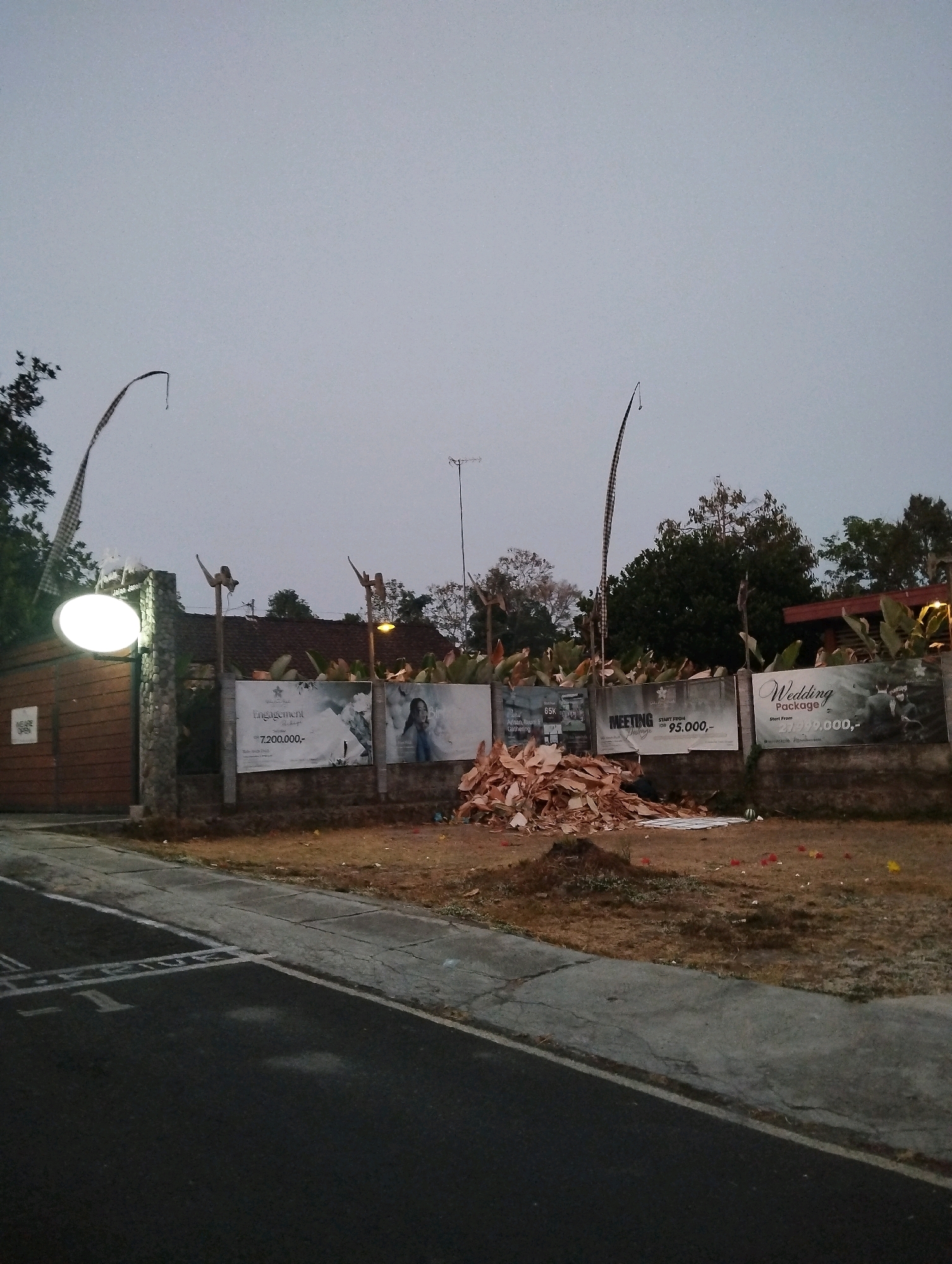 The image shows a view of a property entrance on a cloudy day. The property appears to be in Salatiga, Indonesia, as indicated by the visible text "Salatiga" on a sign. A wooden gate with a stone pillar and a lit oval sign is on the left. Adjacent to the gate, a white banner displays "engagement" with a price of "7,200,000,-". Further to the right, a banner reads "MEETING" with a price of "95.000,-". Another banner on the far right advertises "Wedding Package" starting from "2,999,000,-". In front of the banners, a pile of dry leaves and debris is visible. The ground is covered with dry grass and some scattered red and yellow flowers. A paved road with white markings runs along the bottom of the image. There are no people visible in the scene.