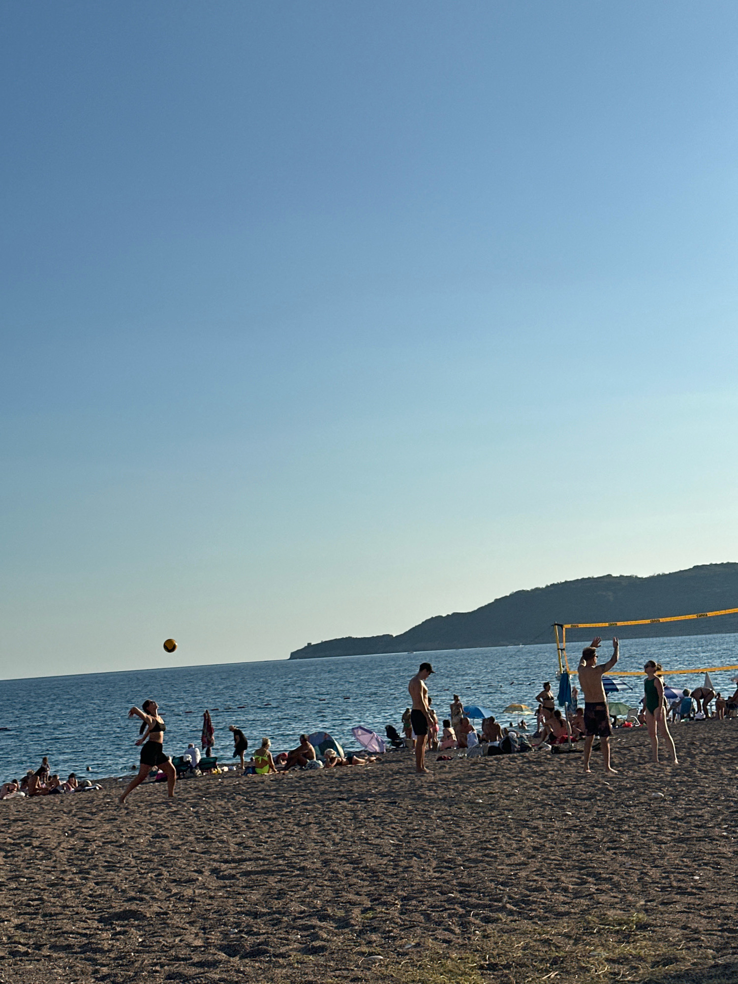 This image captures a sunny day at a sandy beach in Boreti, Montenegro, with a clear blue sky overhead. The foreground is dominated by the dark sand of the beach, with scattered patches of dry grass. In the middle ground, a beach volleyball game is in progress. Four individuals are visible playing on the sand, with a net erected between them. One person is in the process of serving the ball, which is visible in mid-air. Other people are scattered across the beach, some sitting or lying on towels, others standing or walking. Beach umbrellas provide shade for some of the beachgoers. The background features the calm, blue waters of the Adriatic Sea, with a distant, tree-covered hill forming the horizon line. A few boats can be seen on the water. The overall atmosphere is one of leisure and recreation on a warm day at the coast.