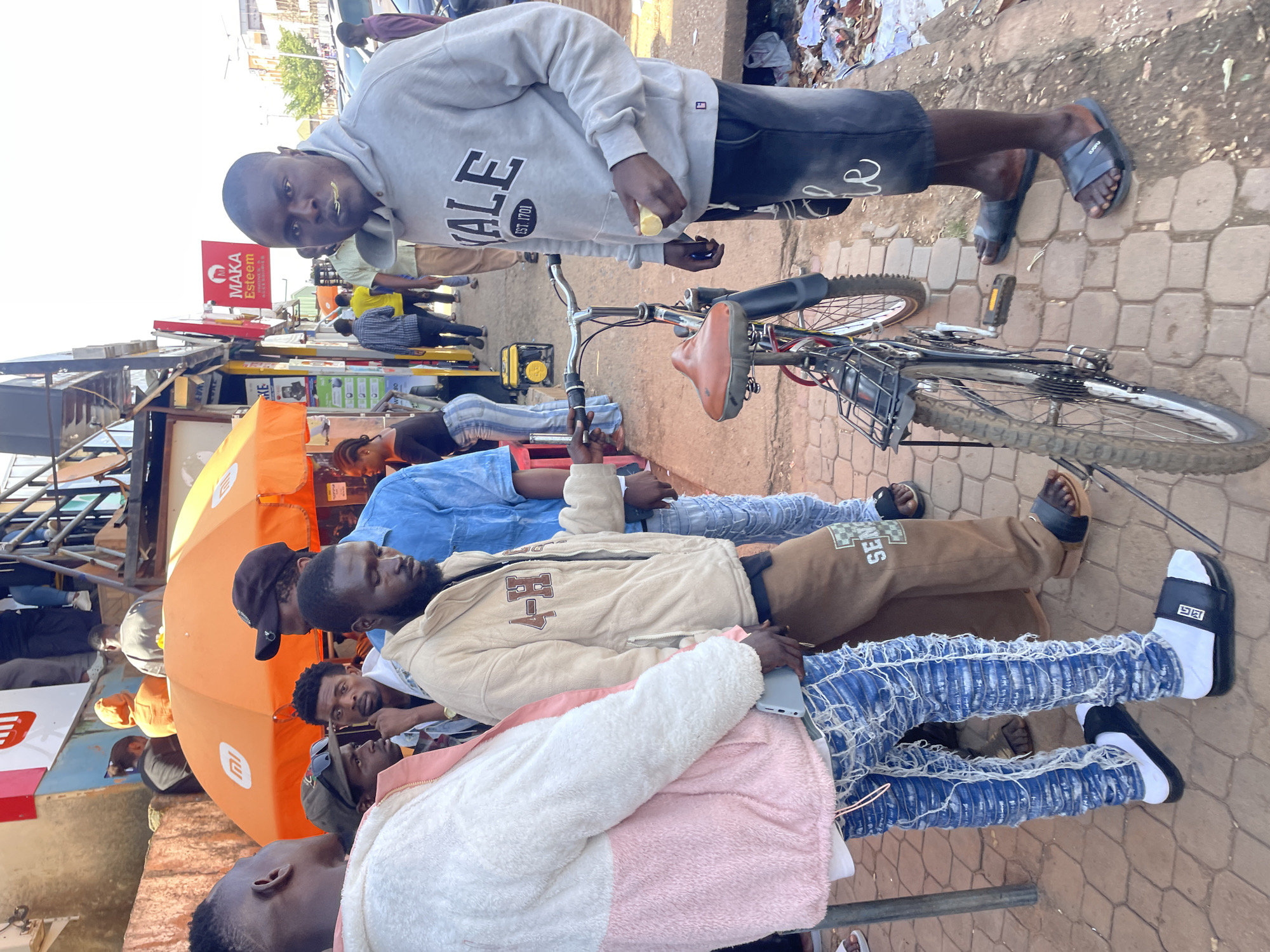 The image depicts a street scene in Jos, Nigeria, focusing on a group of young men standing by a bicycle. The foreground is dominated by the legs of several individuals, one wearing distressed blue jeans with white detailing and black slide sandals over white socks, another in brown sweatpants with a checkered logo on the leg, and a third in dark shorts and black slide sandals. A bicycle with a brown seat and a black frame stands between them.
In the midground, a man wearing a grey hooded sweatshirt with "YALE EST. 1701" printed on it is in the process of putting a piece of yellow fruit into his mouth. He is facing the camera. To his left, another man in a tan jacket with "4-H" printed on the sleeve, dark pants, and sandals is looking down. Further left, a man in a blue denim shirt and light-colored pants is visible. Behind them, an orange umbrella provides shade, with various signs and structures suggesting a market or commercial area. In the background, more people are visible, along with buildings and cars, indicating an urban setting. The ground is paved with interlocking bricks and concrete.