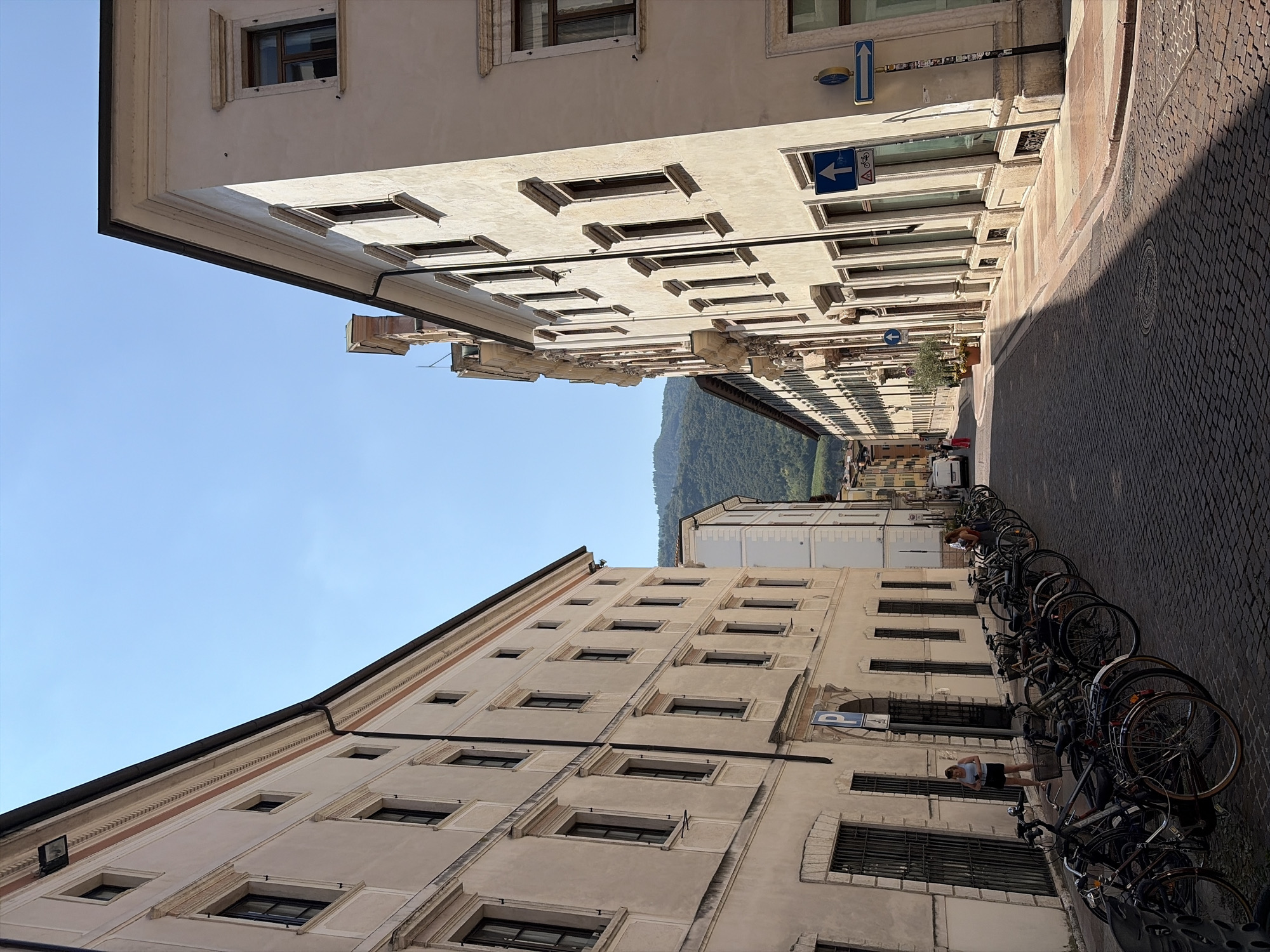 The image depicts a street scene in Trento, Italy, likely during daylight hours. Tall, pale buildings line both sides of a cobblestone street, creating a sense of an urban canyon. The architecture suggests historical significance, with numerous windows and decorative elements on the building facades. A row of parked bicycles occupies the left side of the street, suggesting a common mode of transportation in the area. Two women are present in the scene. One woman, standing near the parked bicycles on the left, is wearing a light-colored top and dark shorts. The second woman, positioned further down the street on the right, is wearing a light-colored top and dark pants. Signs are visible, including directional arrows and a bicycle symbol, indicating traffic regulations or pedestrian/cyclist pathways. In the background, a hillside covered in vegetation is visible, with a clear blue sky overhead. The sunlight casts shadows across the cobblestone street, indicating the sun's position.